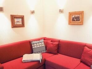 A red sofa with a patterned throw pillow and a blanket, two framed pictures on the wall, and a light fixture. - Chesa Montana - Top 101 (Maria Alm am Steinernen Meer)