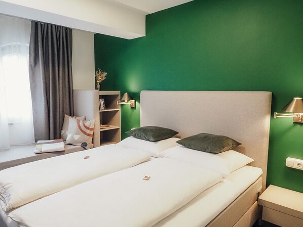 Ein Hotelzimmer mit zwei Betten, einer grünen Wand, einem Bücherregal und einem Fenster mit Vorhängen.