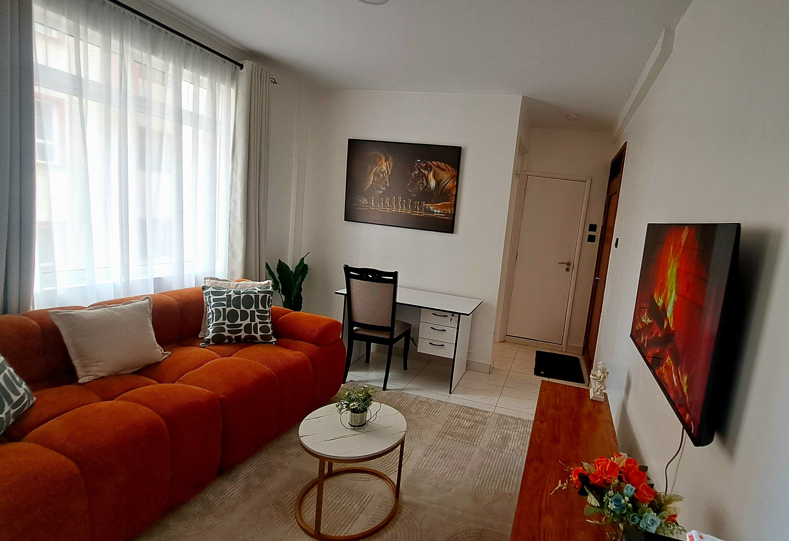 Un salon avec un canapé, une petite table ronde, un bureau et une chaise. Il y a un tableau au mur et une porte au fond.
