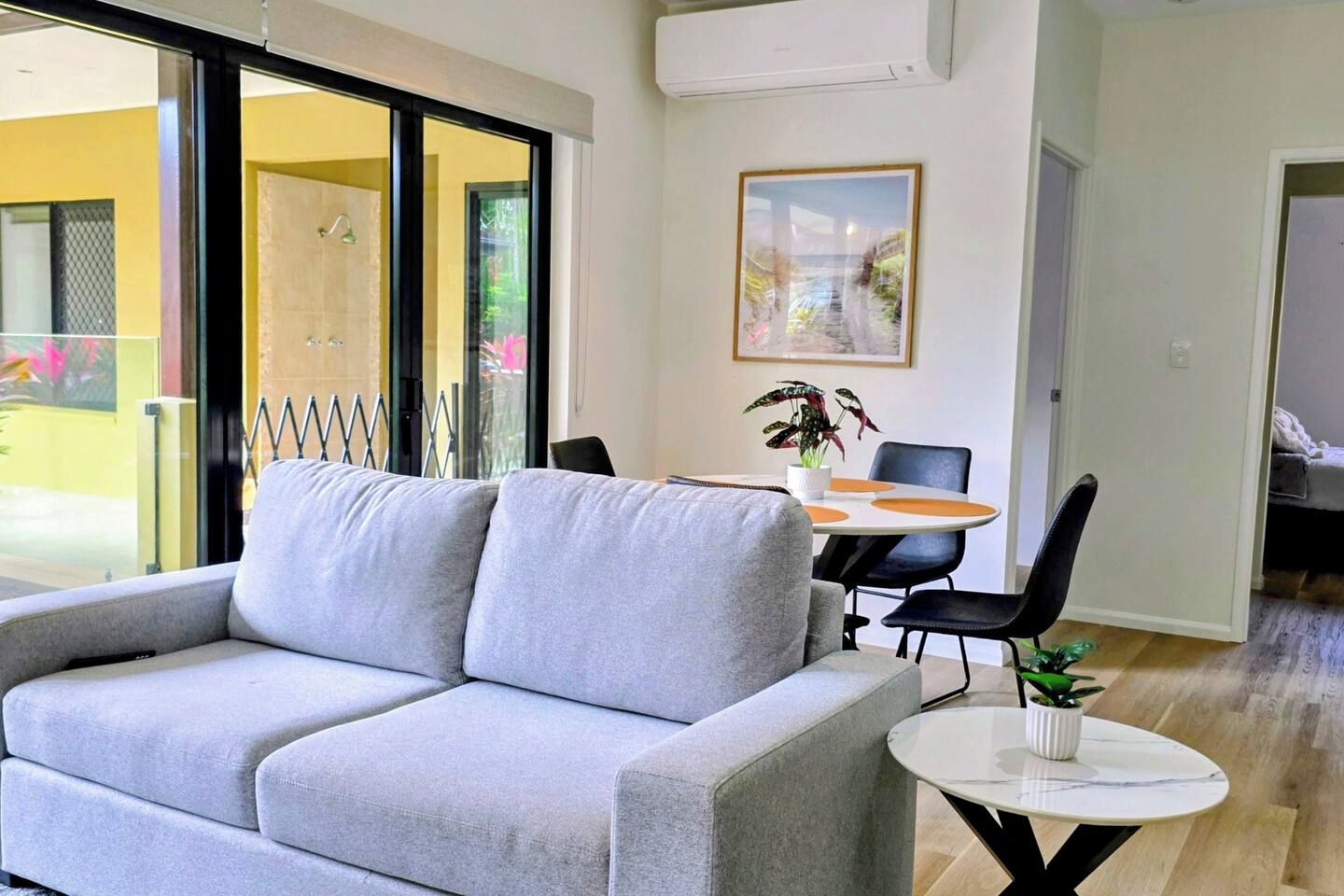 A modern living room with a grey sofa, a dining table with chairs, a small round table with a plant, and a large window with a view of the outdoors.