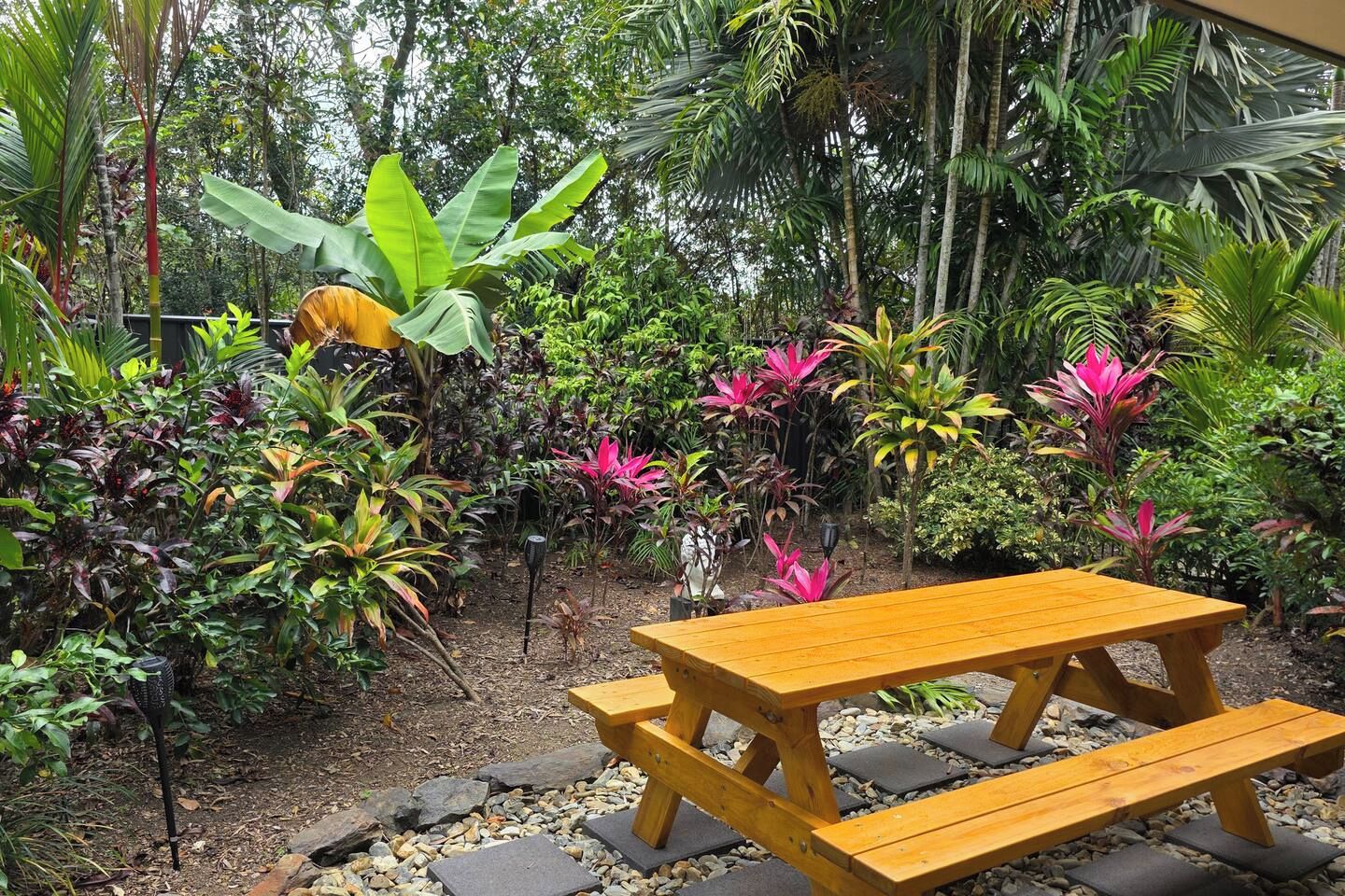 A wooden picnic table in a garden with various tropical plants and a stone pathway.