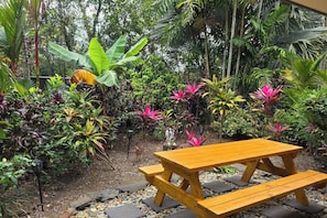 A wooden picnic table in a garden with various tropical plants and a stone pathway.