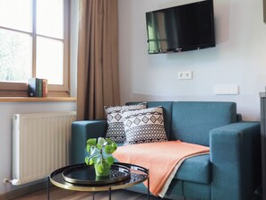 Ett vardagsrum med en blå soffa, mönstrade kuddar, ett litet runt bord med en växt, en tv som är monterad på väggen och ett fönster med gardiner.