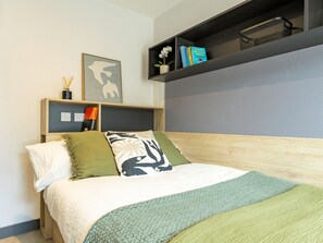 Ein ordentlich bezogenes Bett mit grüner Decke und Kissen, ein kleines Regal mit Büchern und einer Blume sowie eine Wand mit einem gerahmten Bild.