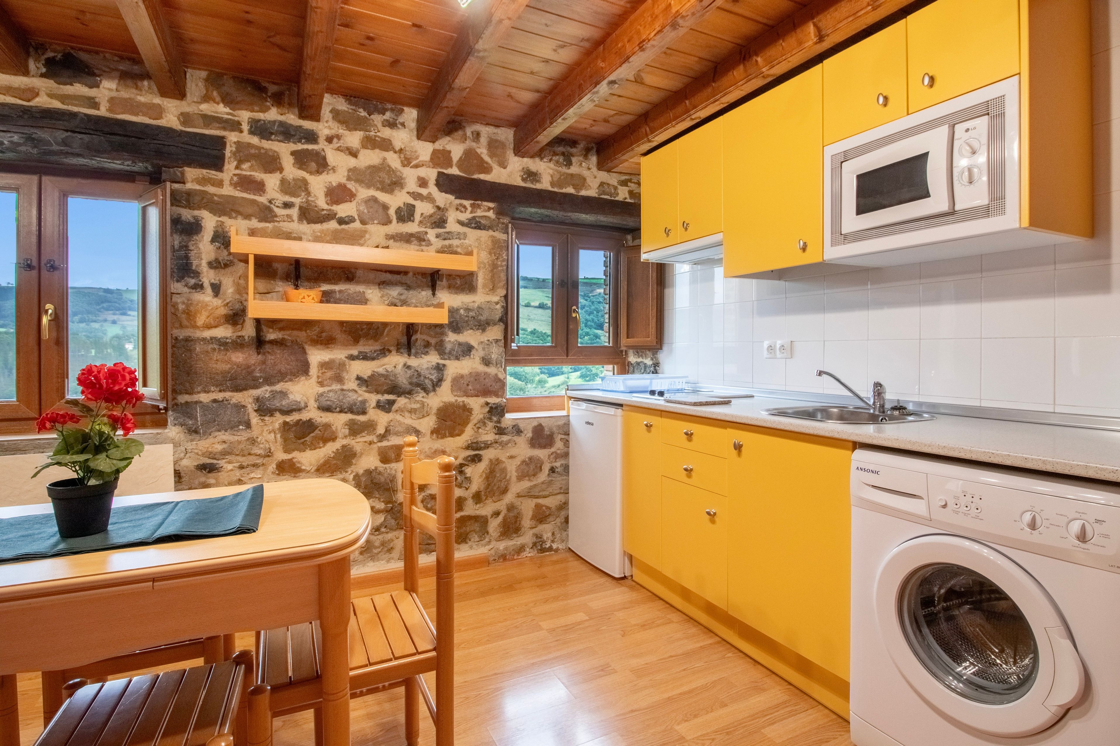 Een keuken met stenen muren, een houten plafond, gele kastjes, een wasmachine en een eethoek met een tafel en stoelen.