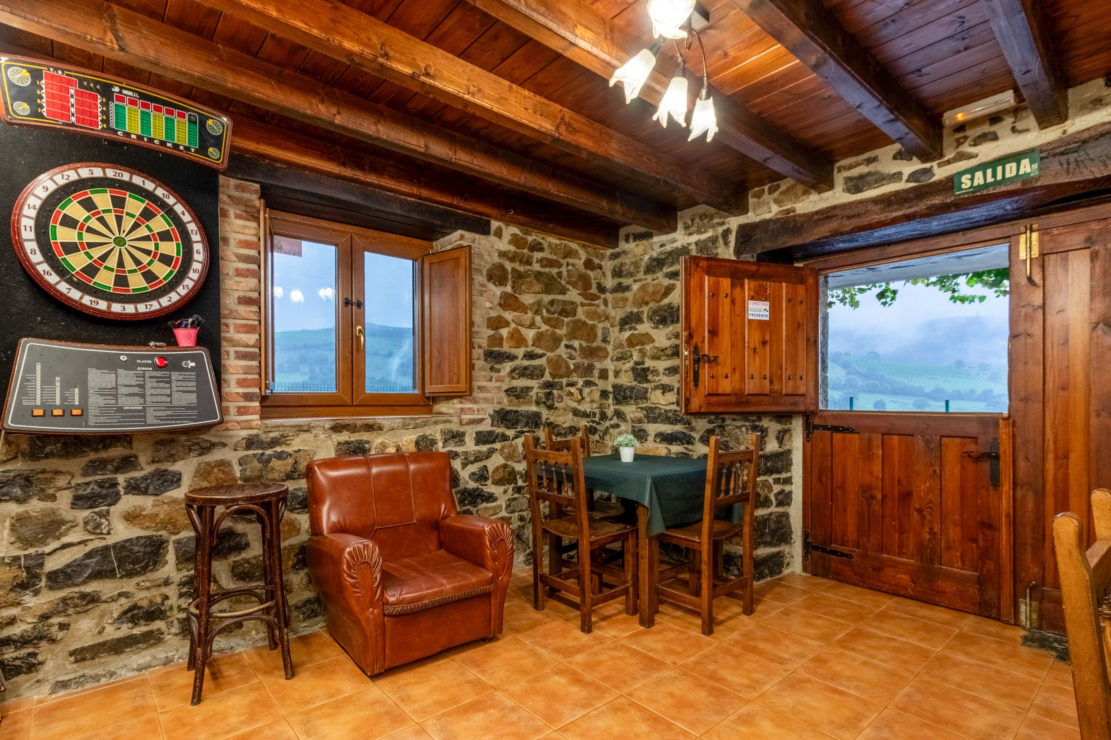 A room with a dartboard, a wooden chair, a table with chairs, a stone wall, and a view through a window.