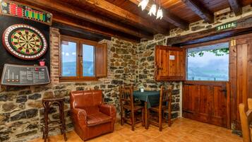 A room with a dartboard, a wooden chair, a table with chairs, a stone wall, and a view through a window.