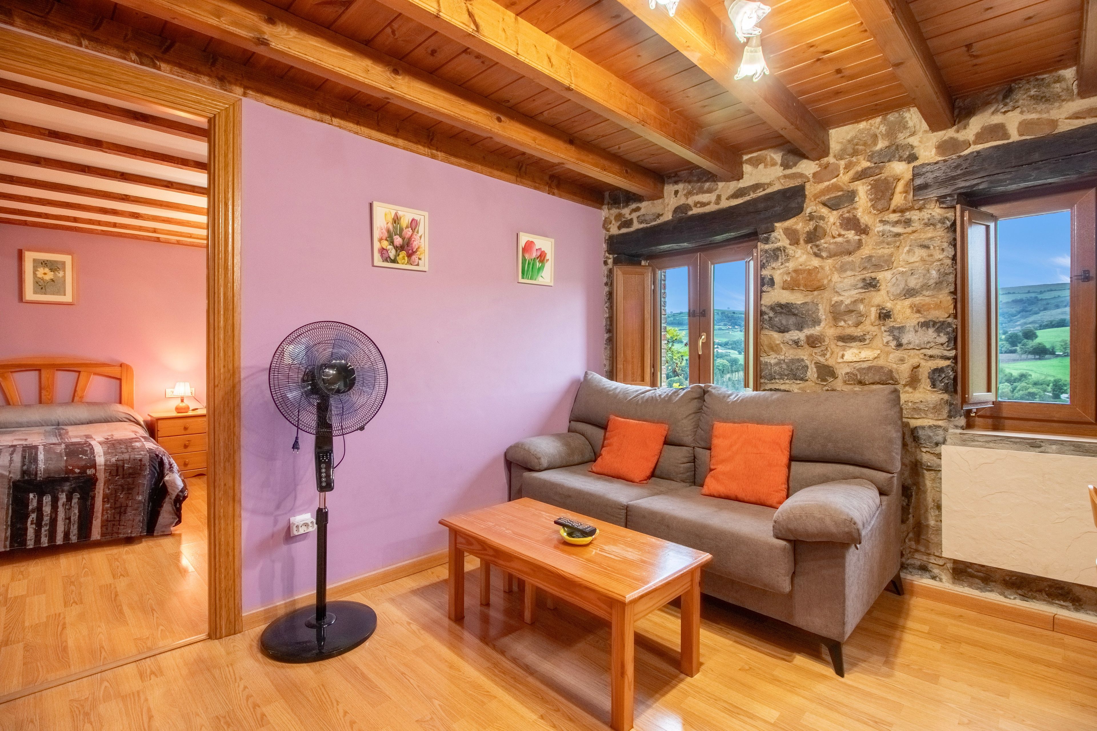 Quarto aconchegante com parede de pedra, teto de madeira, sofá com almofadas laranja, mesa de madeira e um ventilador.
