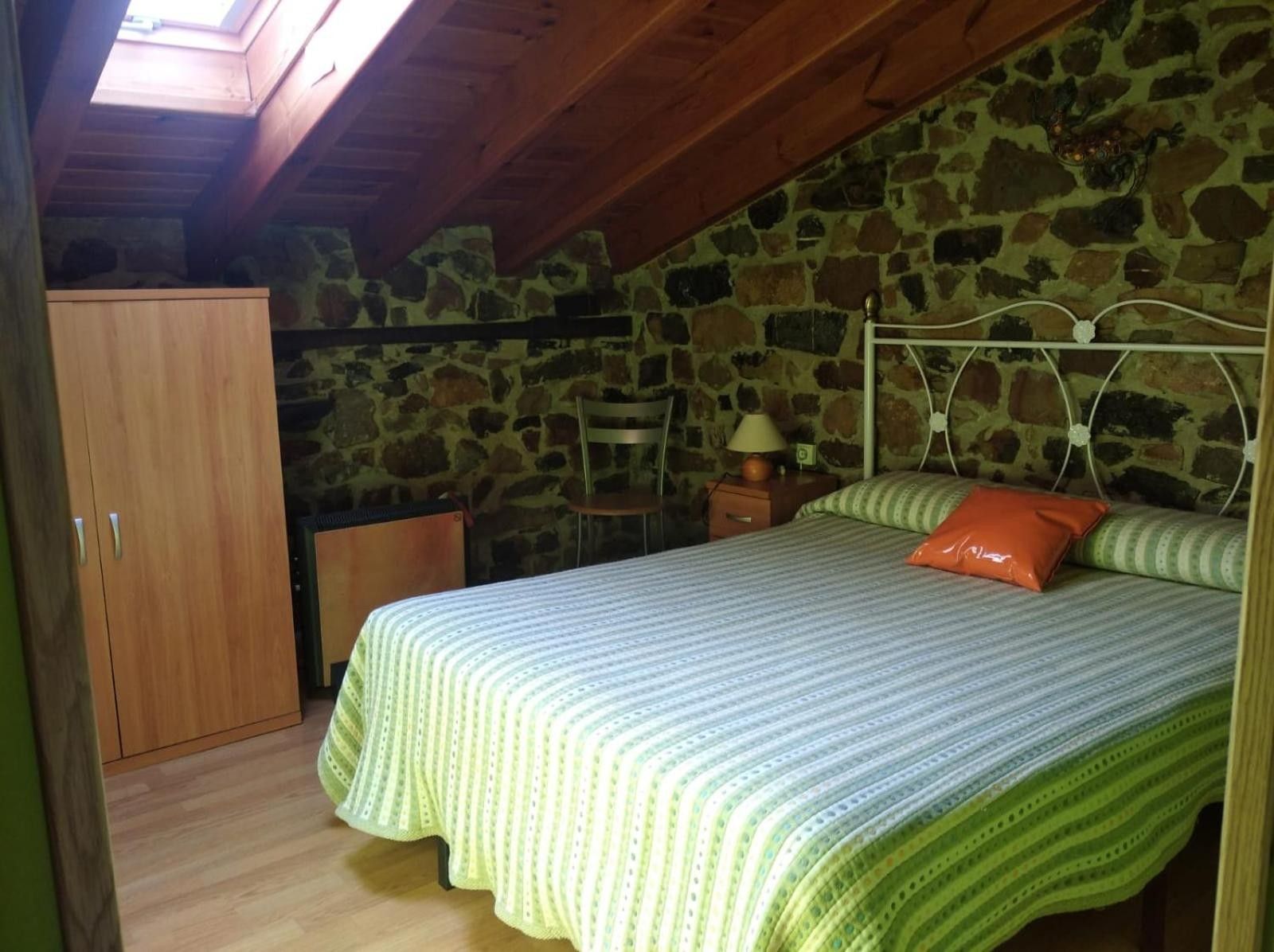 Un dormitorio con pared de piedra, techo de madera, cama con ropa de cama verde, mesita de noche y armario.