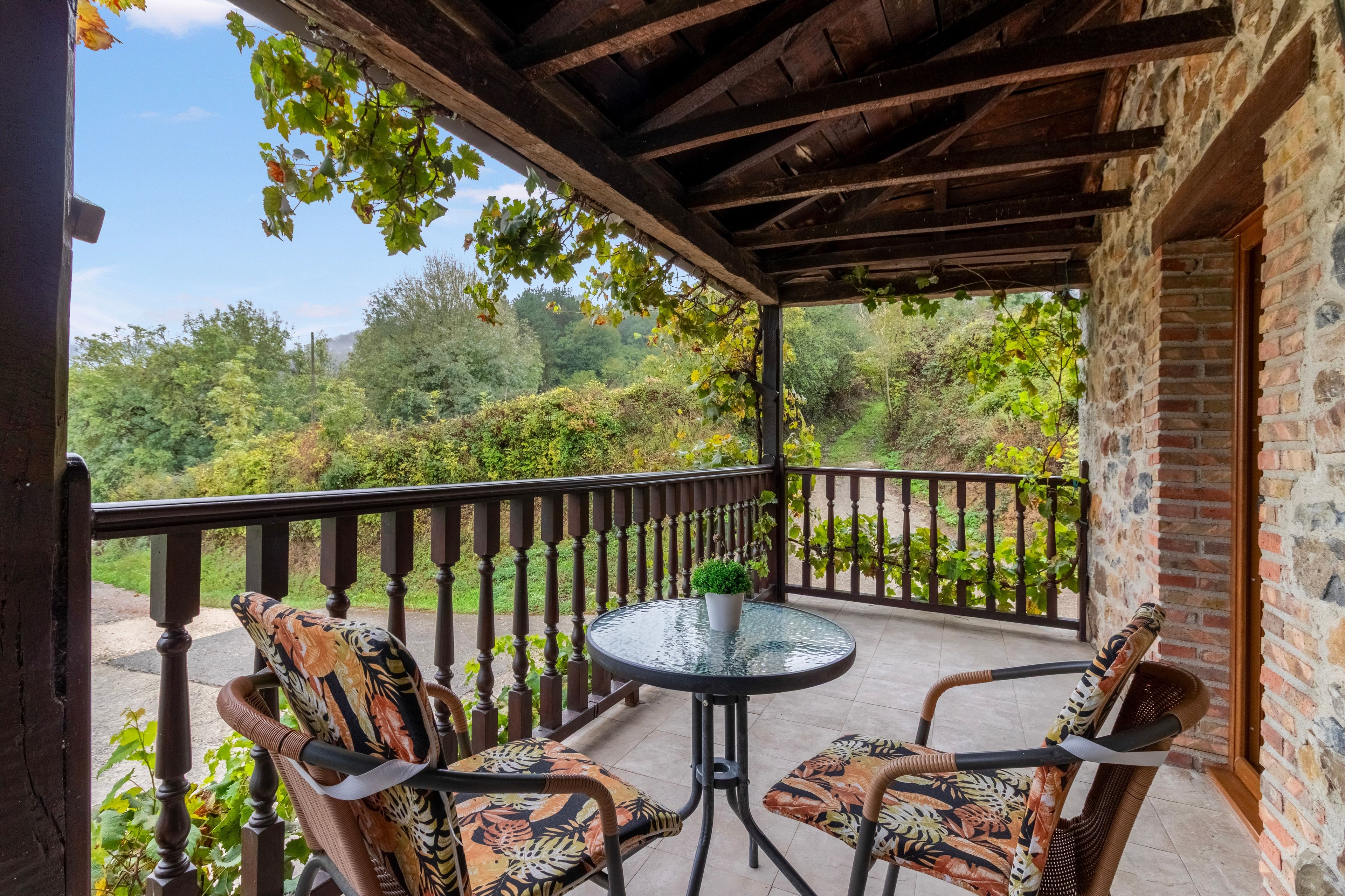 A covered outdoor seating area with two chairs and a table, overlooking a lush garden and a stone building.