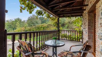 A covered outdoor seating area with two chairs and a table, overlooking a lush garden and a stone building.