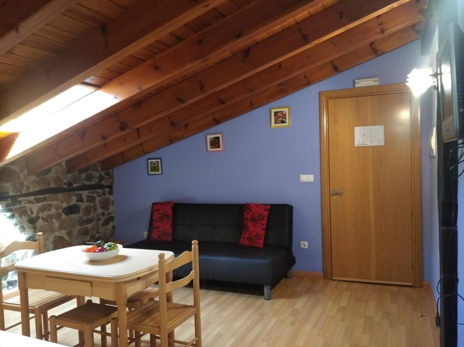Una habitación con techo de madera, pared de piedra, sofá azul con cojines rojos, mesa de madera con un cuenco de fruta y una puerta de madera.