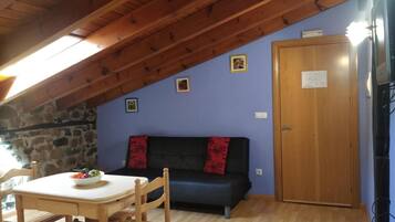 Una habitación con techo de madera, pared de piedra, sofá azul con cojines rojos, mesa de madera con un cuenco de fruta y una puerta de madera.