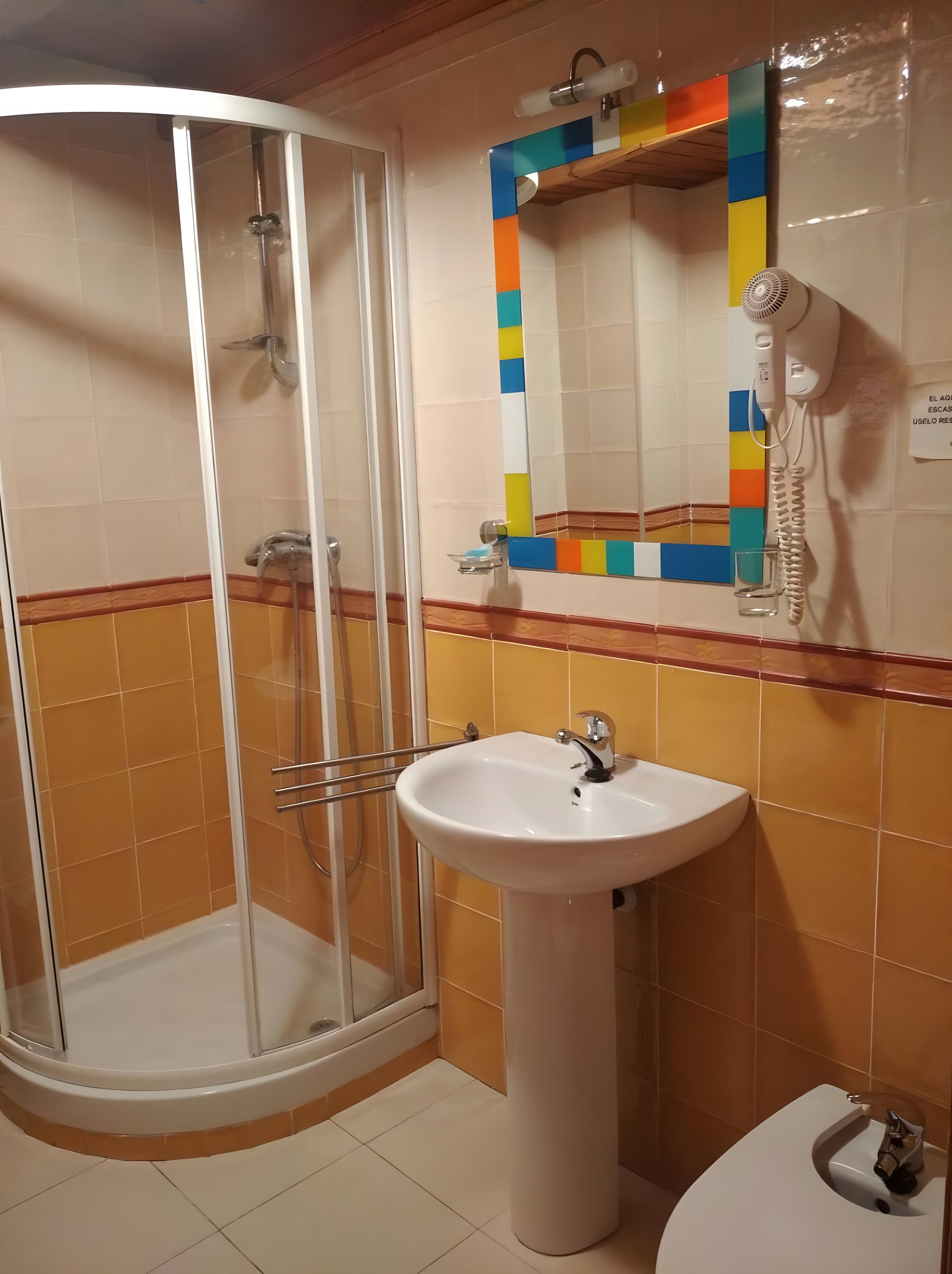 Un baño con ducha envuelta en vidrio, lavamanos sobre pedestal y espejo con detalle de cerámica de colores.