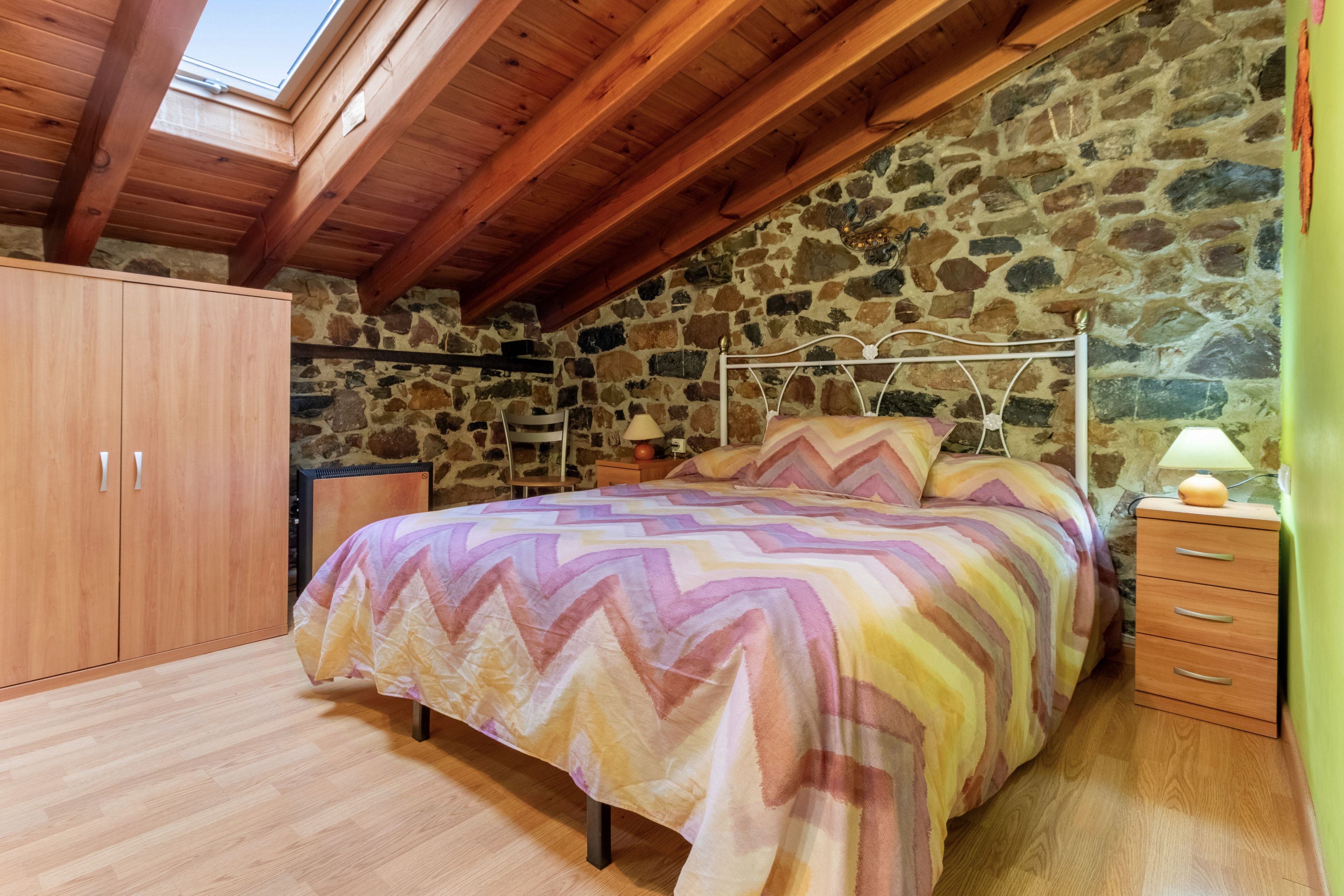 Een slaapkamer met een stenen muur, houten balken in het plafond, een bed met een bedspread met patroon, een nachtkastje en een kledingkast.