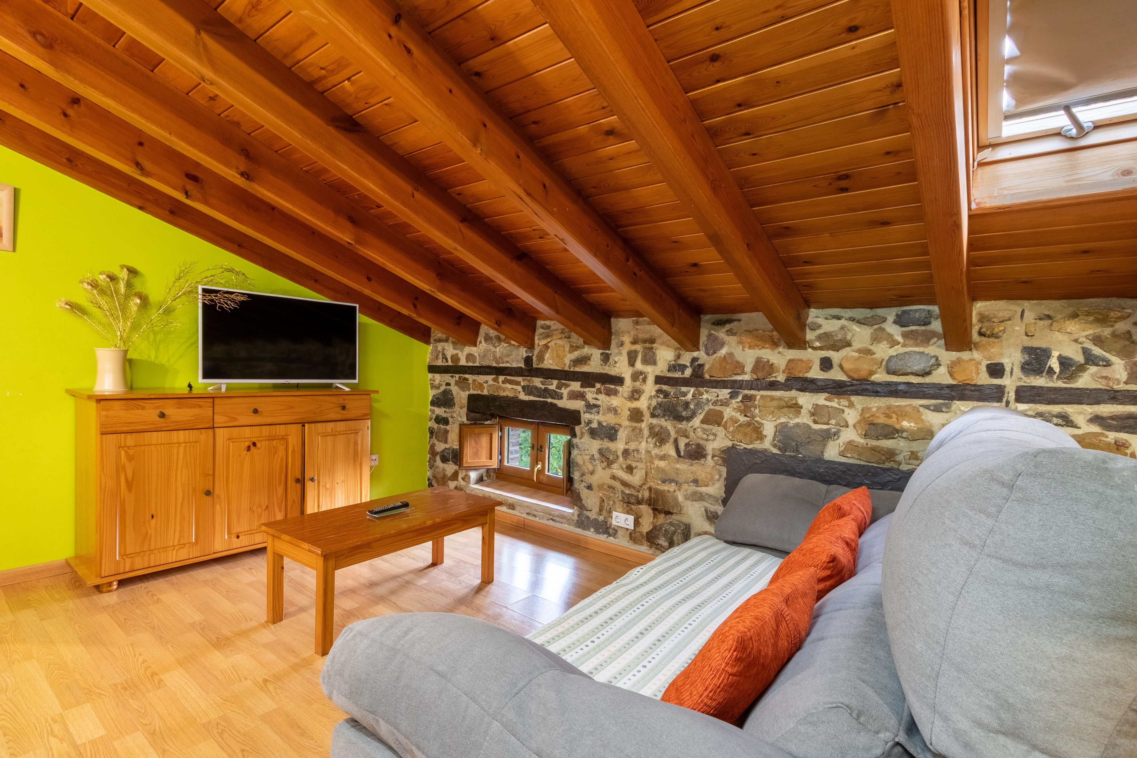 A cozy living room with a stone wall, wooden ceiling beams, a sofa, a coffee table, and a television on a cabinet.