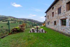 Un bâtiment en pierre avec un toit en pierre, un jardin clos avec de la pelouse, et une vue sur des collines en arrière-plan.
