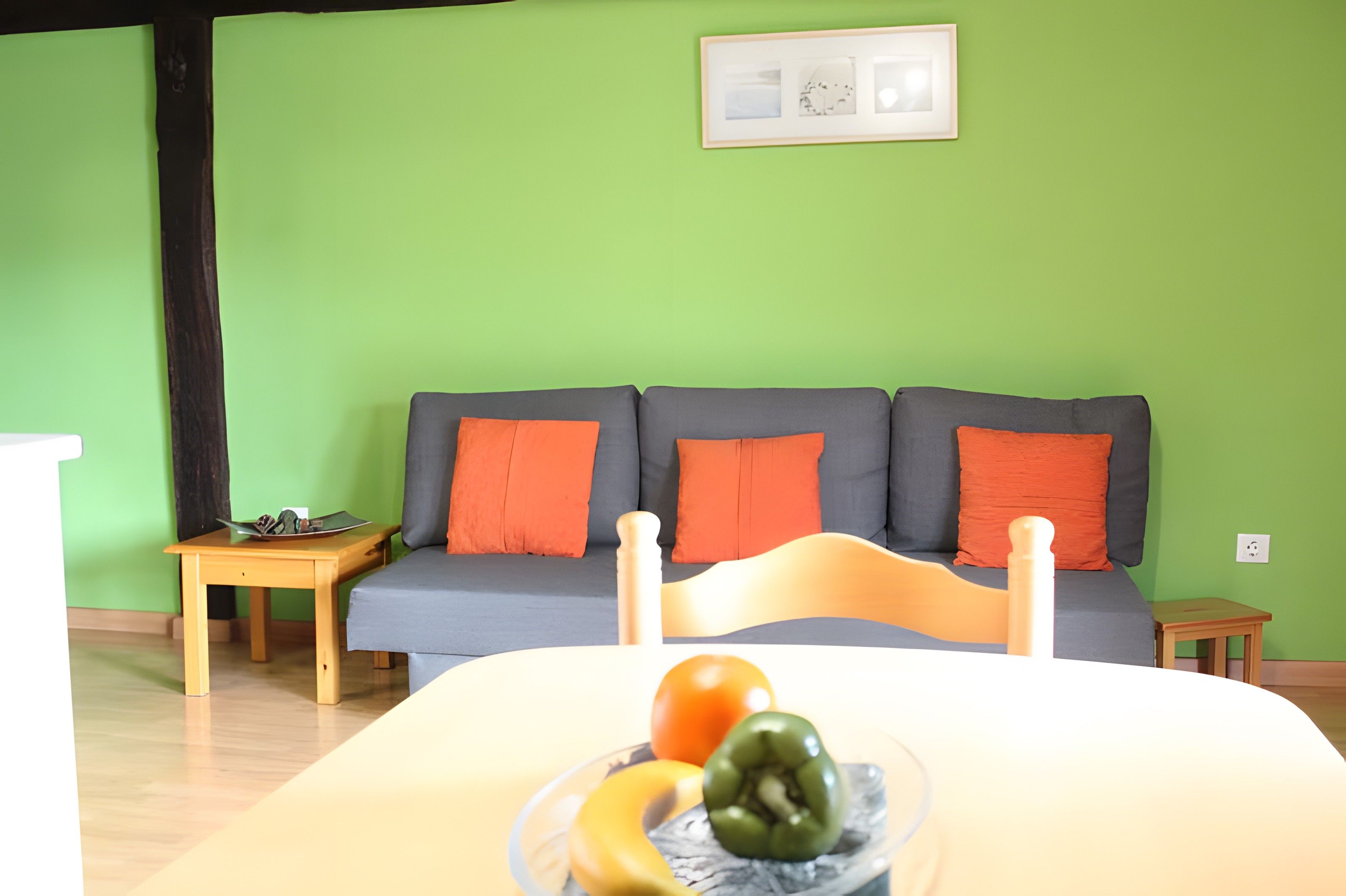 Una habitación con una pared verde, un sofá gris con cojines naranjas, una mesa de madera y una cama con un frutero encima.