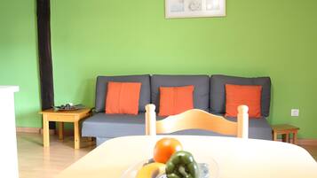 Una habitación con una pared verde, un sofá gris con cojines naranjas, una mesa de madera y una cama con un frutero encima.