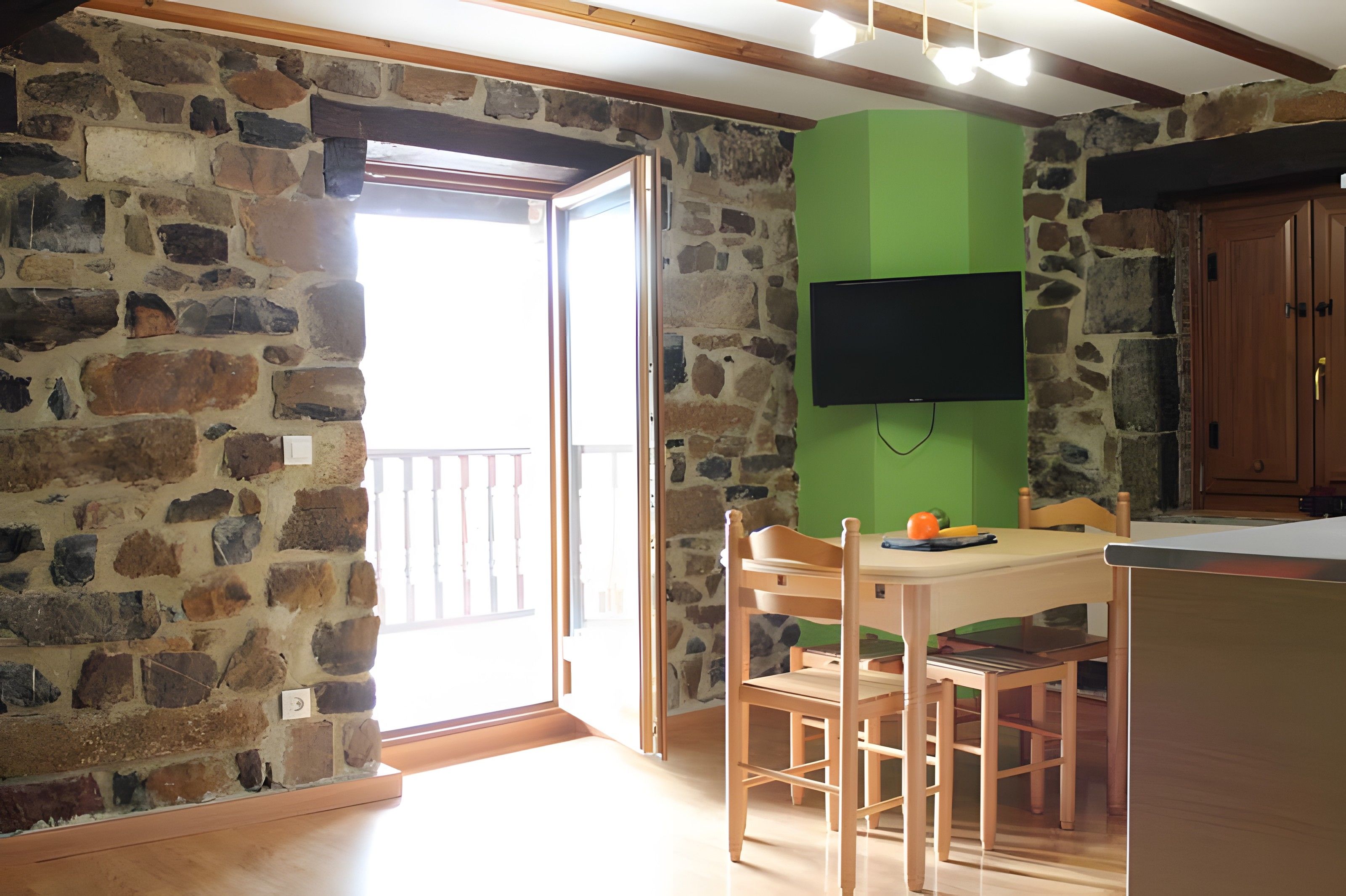 Una habitación con pared de piedra, puerta de madera, pared verde, televisor montado en la pared, mesa de madera con sillas y un mueble de madera.