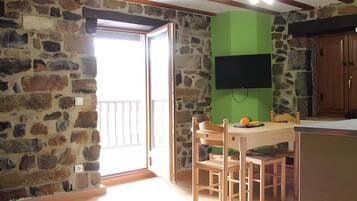 Una habitación con pared de piedra, puerta de madera, pared verde, televisor montado en la pared, mesa de madera con sillas y un mueble de madera.