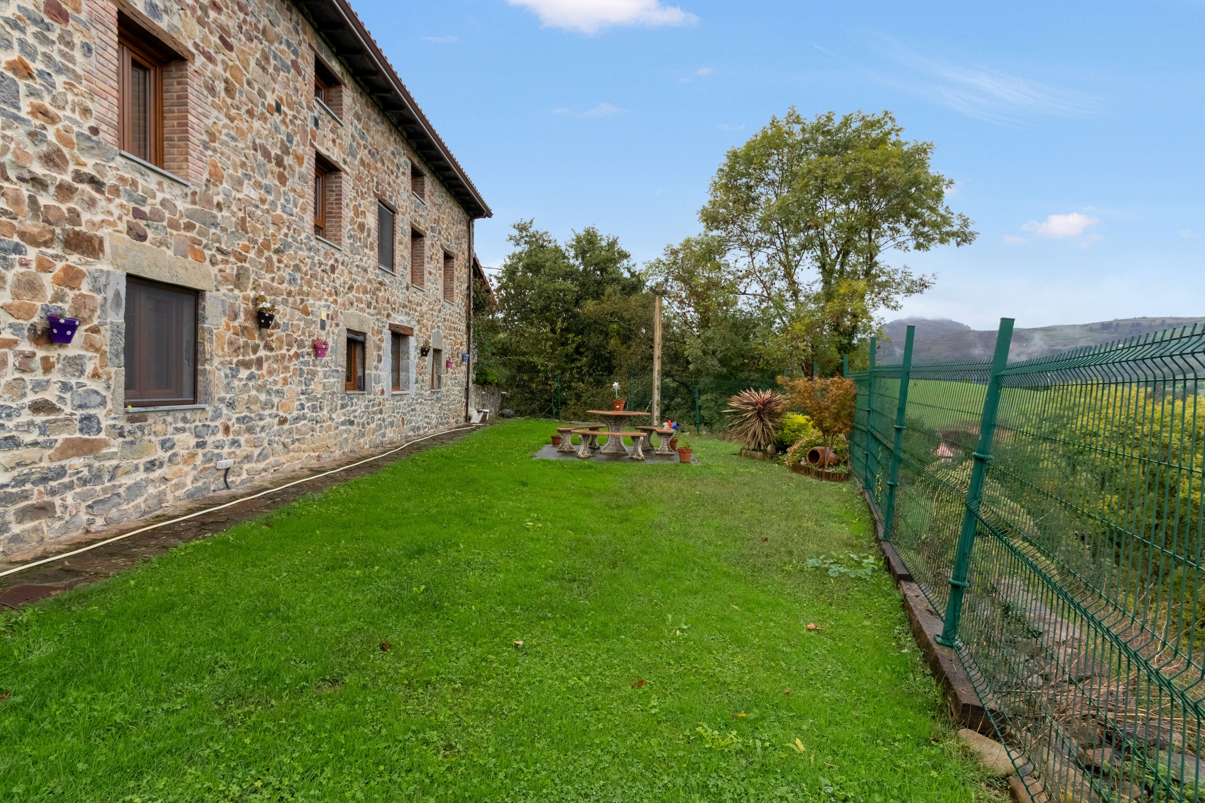 Un edificio in pietra con un cortile erboso, un tavolo e sedie in legno e una recinzione verde.