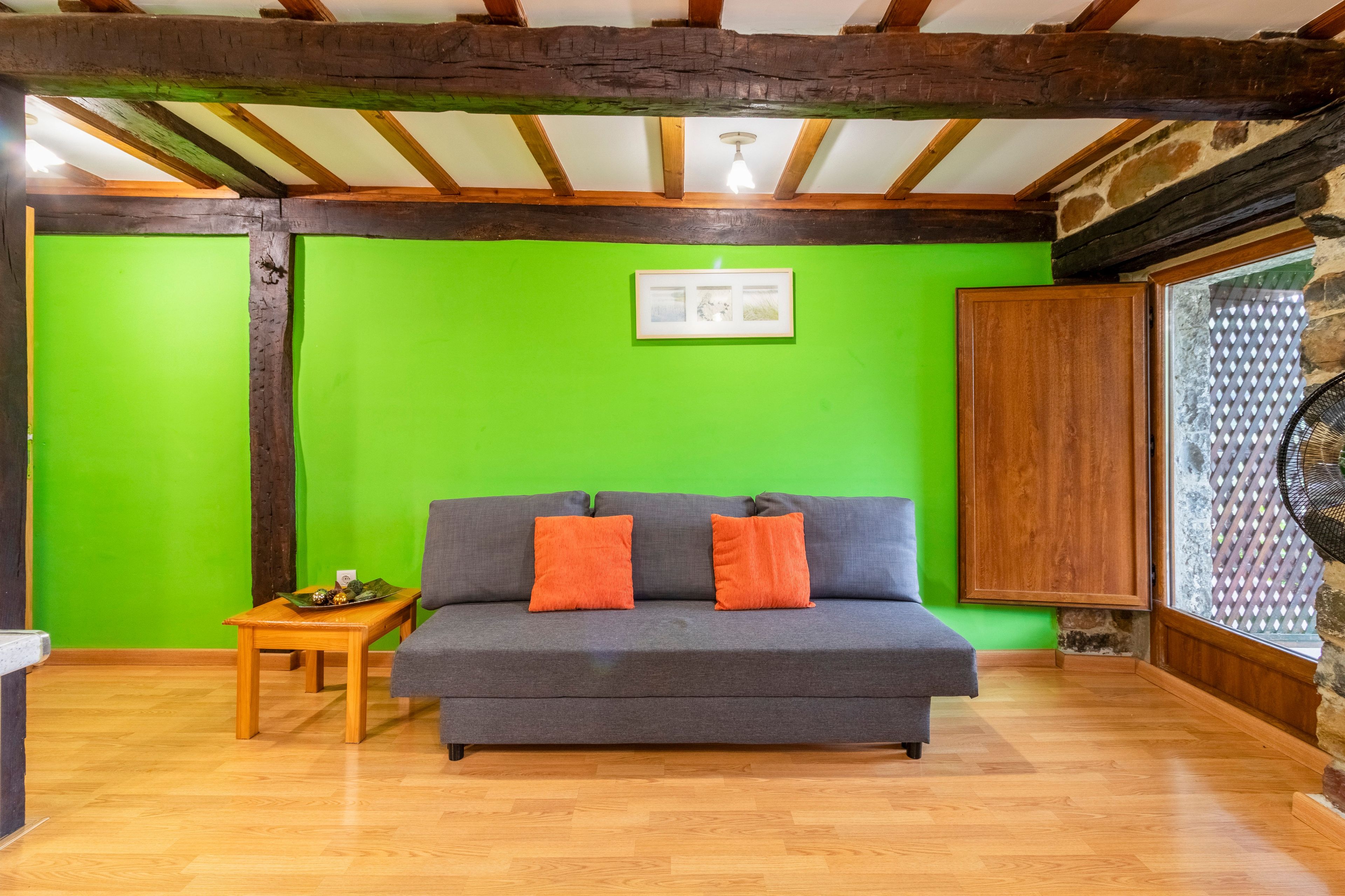 Een kamer met een grijze bank, twee oranje kussens, een klein houten tafeltje met een decoratief object, en een groene wand.