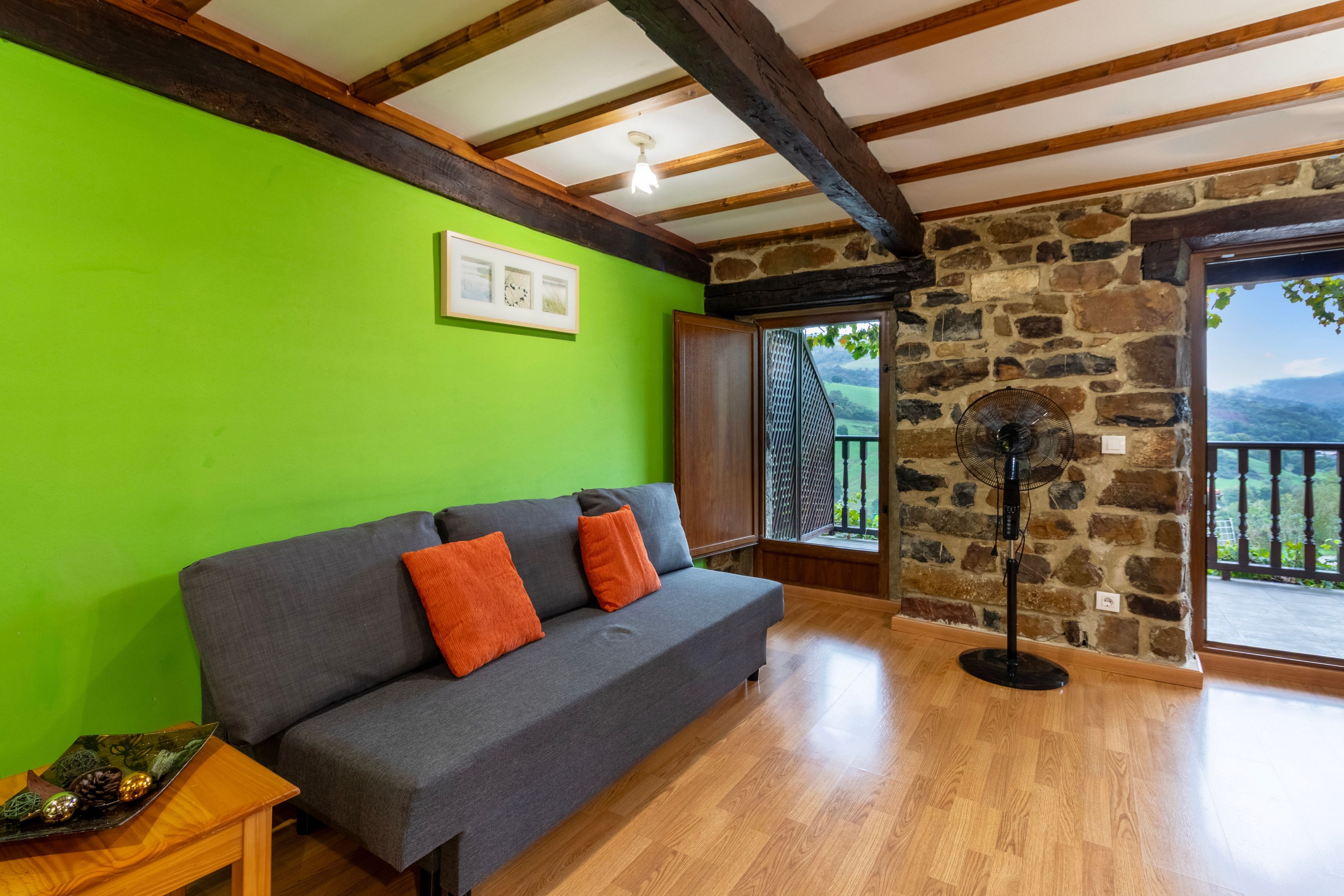 Una habitación con sofá gris, suelo de madera, pared verde y pared de piedra, además de un ventanal con vistas.