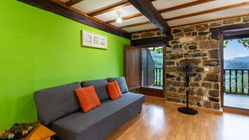 Una habitación con sofá gris, suelo de madera, pared verde y pared de piedra, además de un ventanal con vistas.