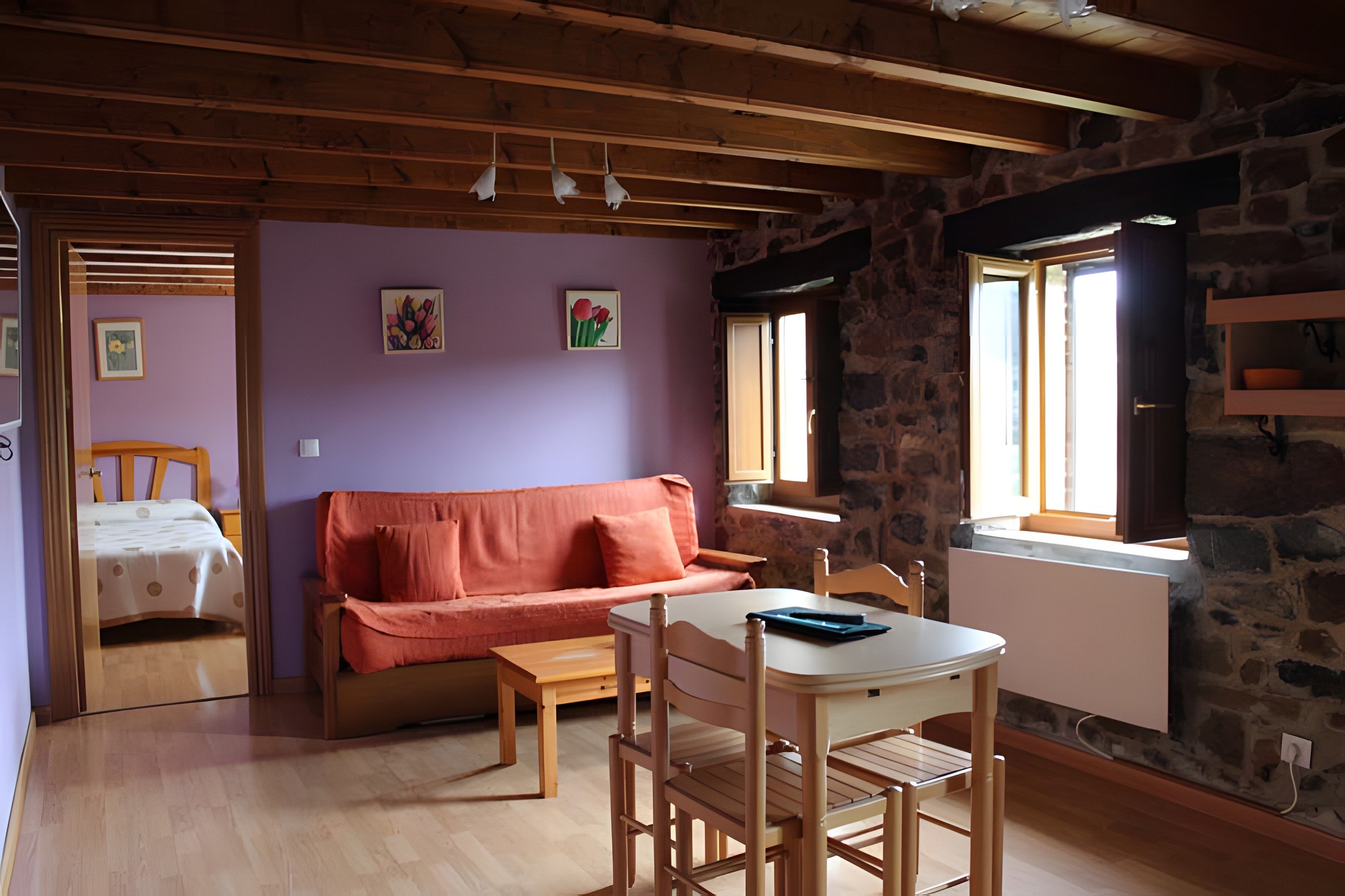 Een gezellige kamer met een houten plafond, een stenen muur, een paarse muur, een houten tafel met stoelen, een bank en een bed met stippenbeddels.