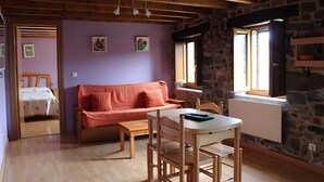 Una habitación acogedora con techo de madera, pared de piedra, pared morada, una mesa de madera con sillas, un sofá y una cama con colcha de lunares.