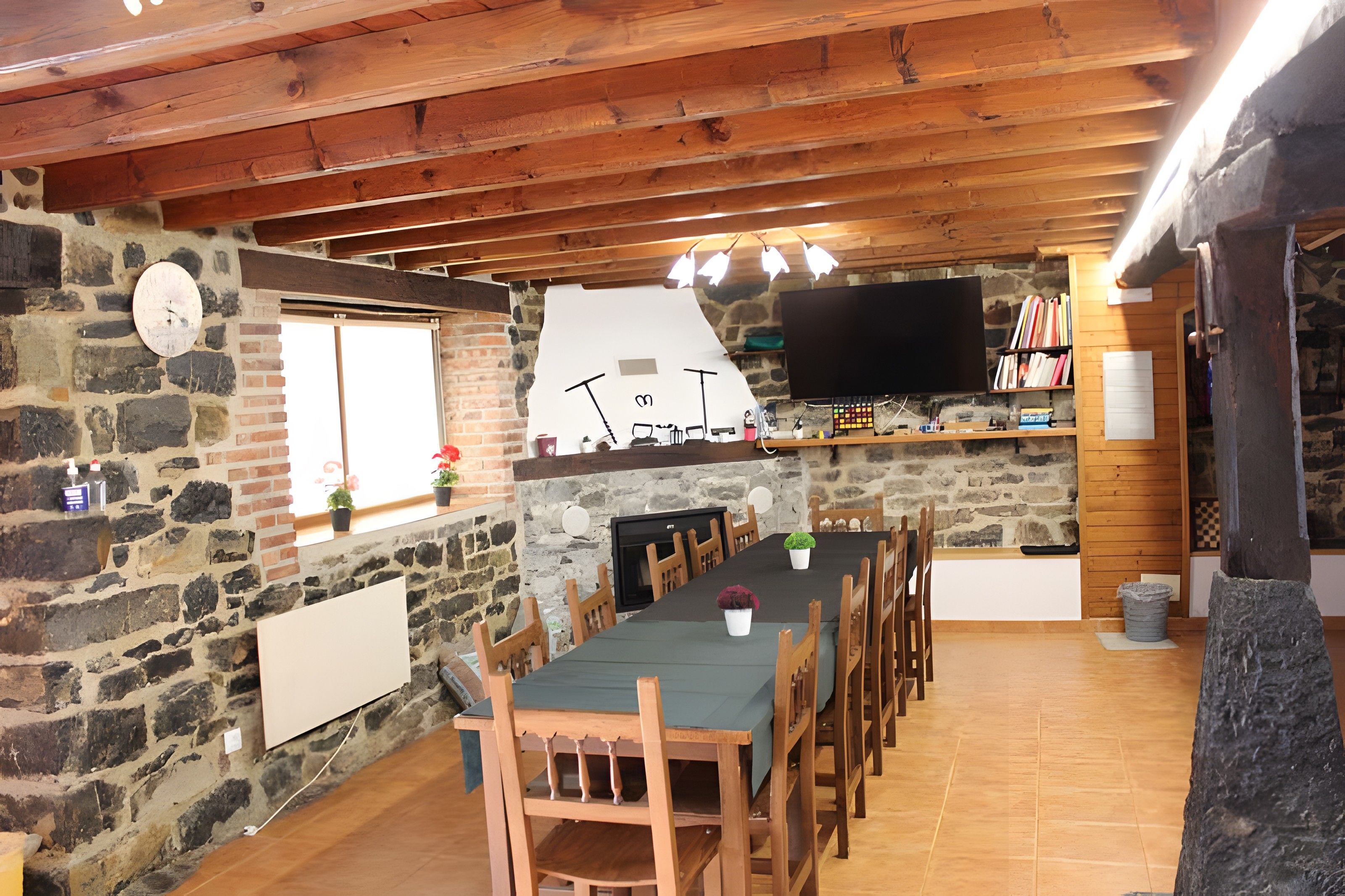 Un coin repas rustique avec un mur en pierre, des poutres en bois au plafond, un téléviseur à écran plat fixé au mur et une longue table entourée de chaises.