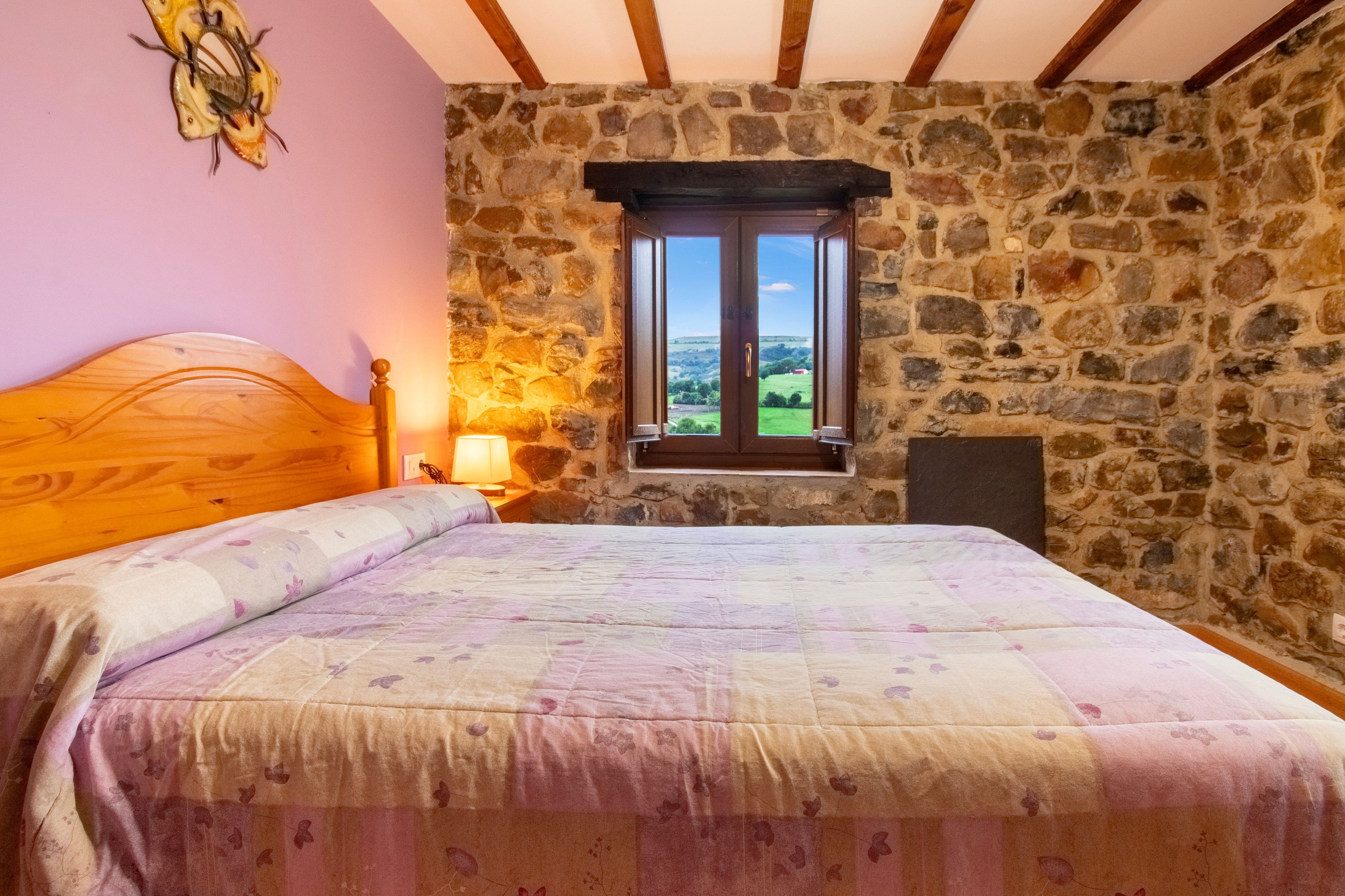 Une chambre avec un mur en pierre, un lit en bois, une fenêtre donnant sur un beau paysage et une table de chevet avec une lampe.