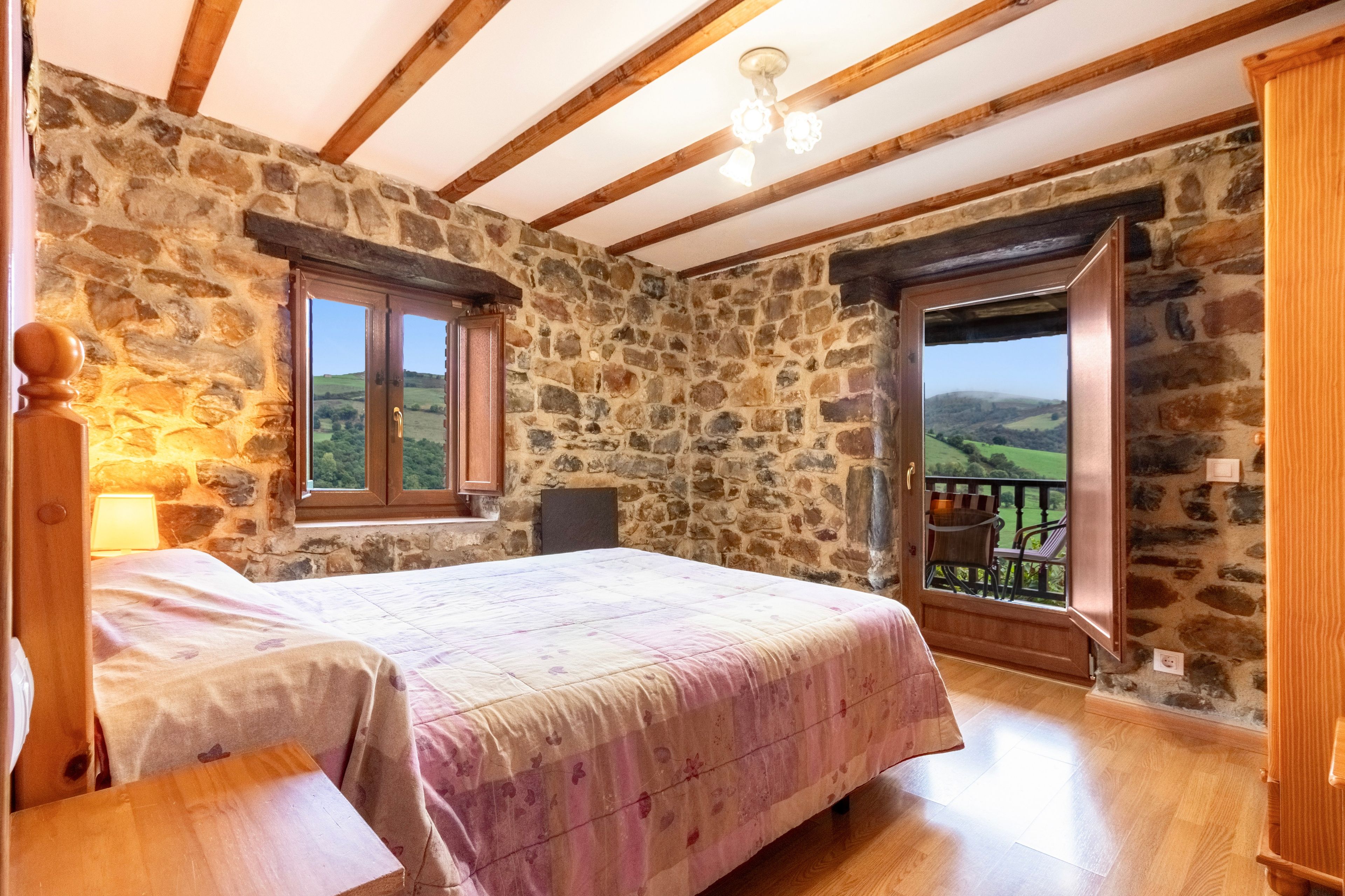 Un dormitorio con pared de piedra, techo de madera, cama con colcha estampada y vista a un paisaje a través de una puerta abierta.