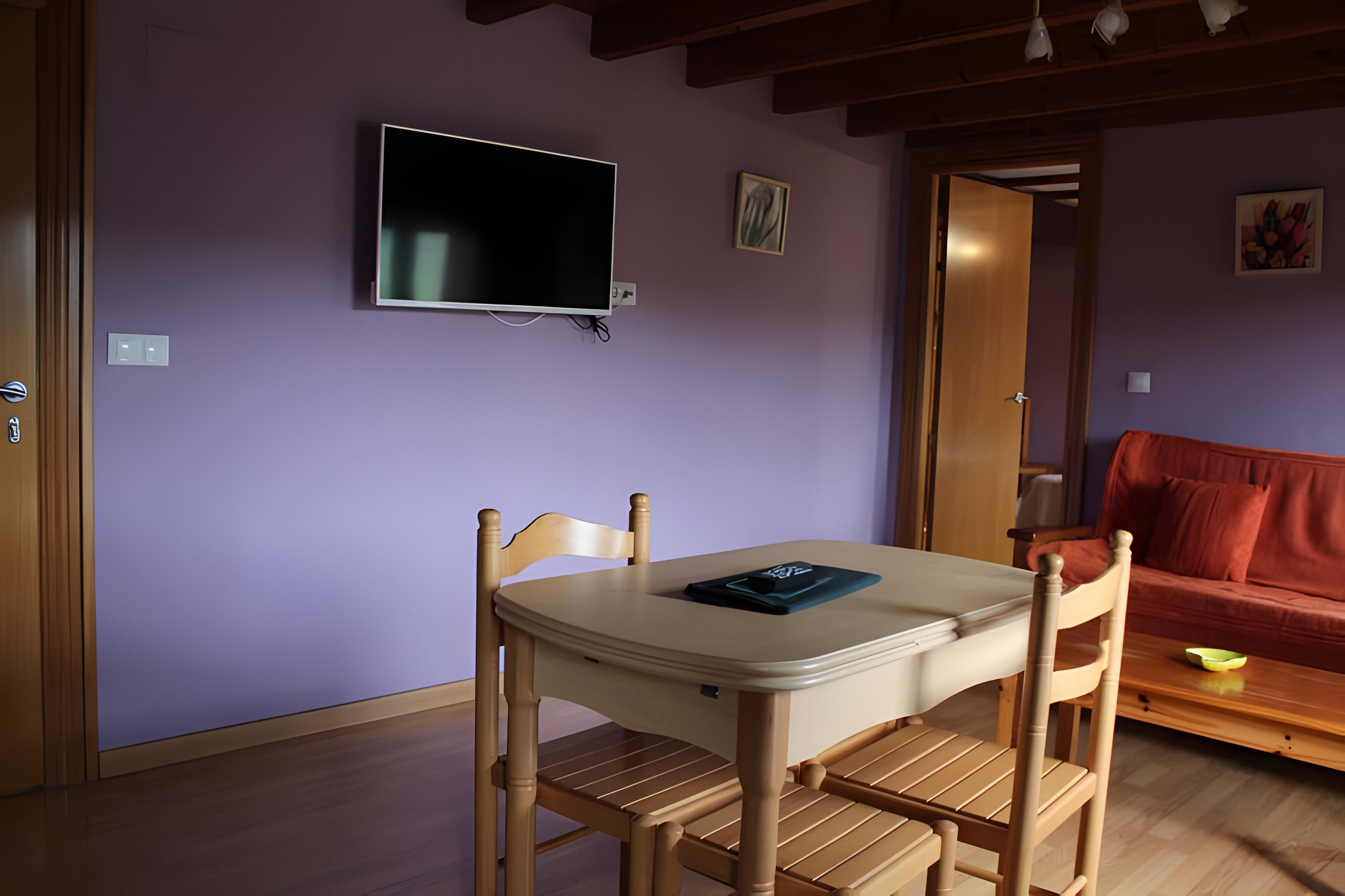 Una habitación con una mesa y sillas, un televisor montado en la pared y una puerta que da a otra habitación.