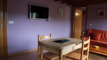 Una habitación con una mesa y sillas, un televisor montado en la pared y una puerta que da a otra habitación.