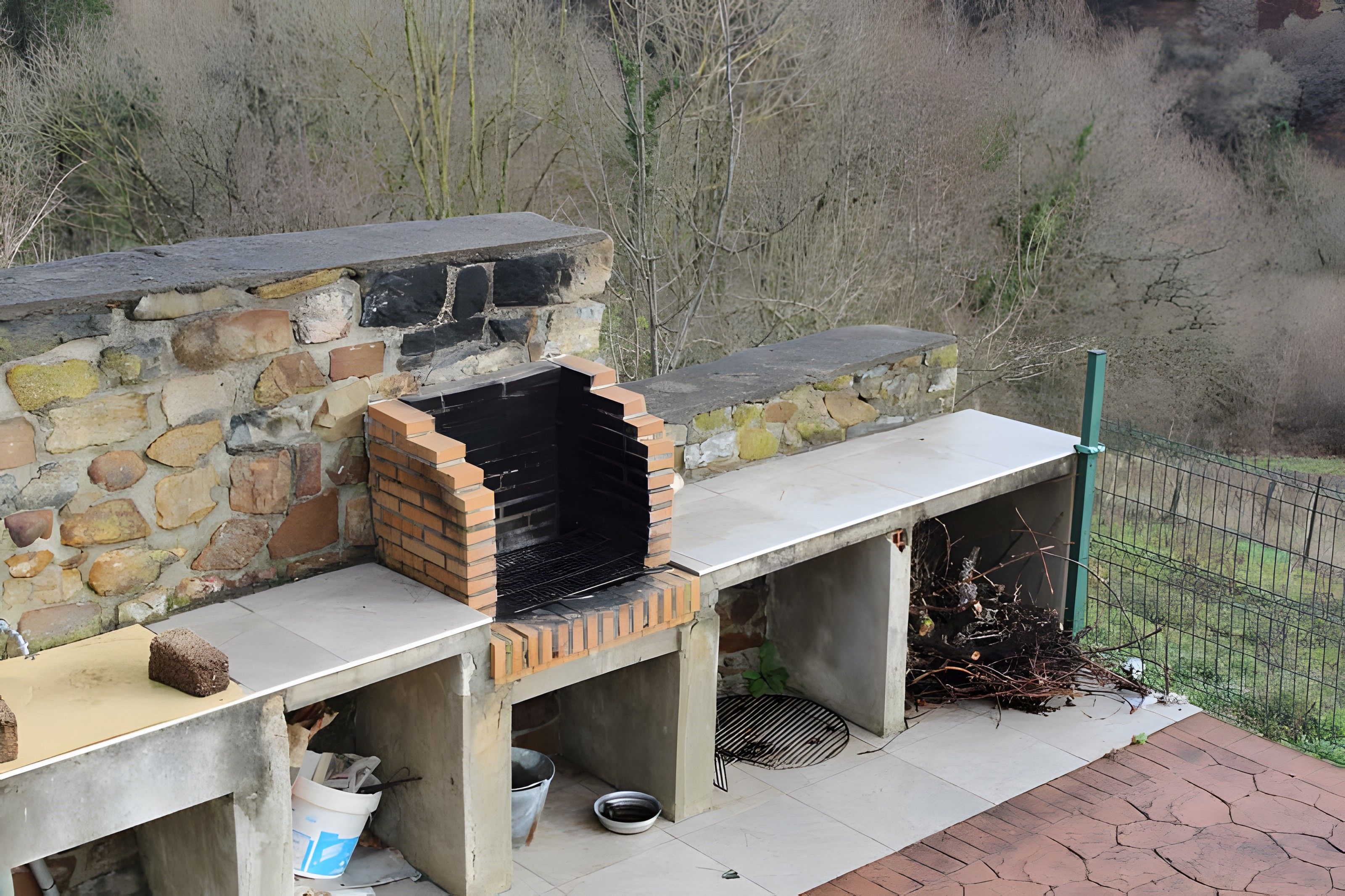 Una zona de cocina al aire libre con una parrilla de piedra y ladrillo, una mesa de concreto y una plancha metálica.