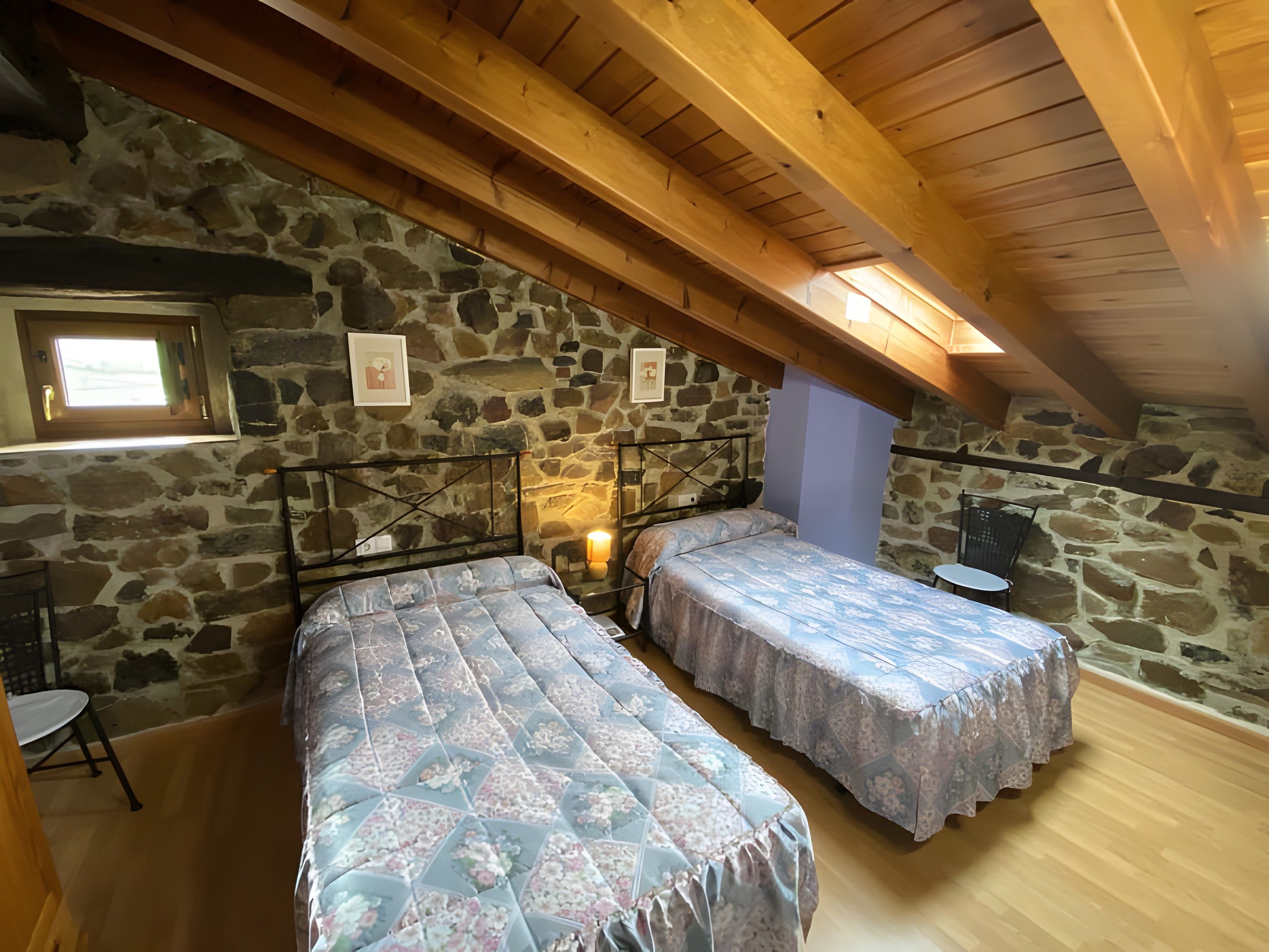 Une chambre avec deux lits, des murs en pierre, des poutres en bois, une petite table et une fenêtre.