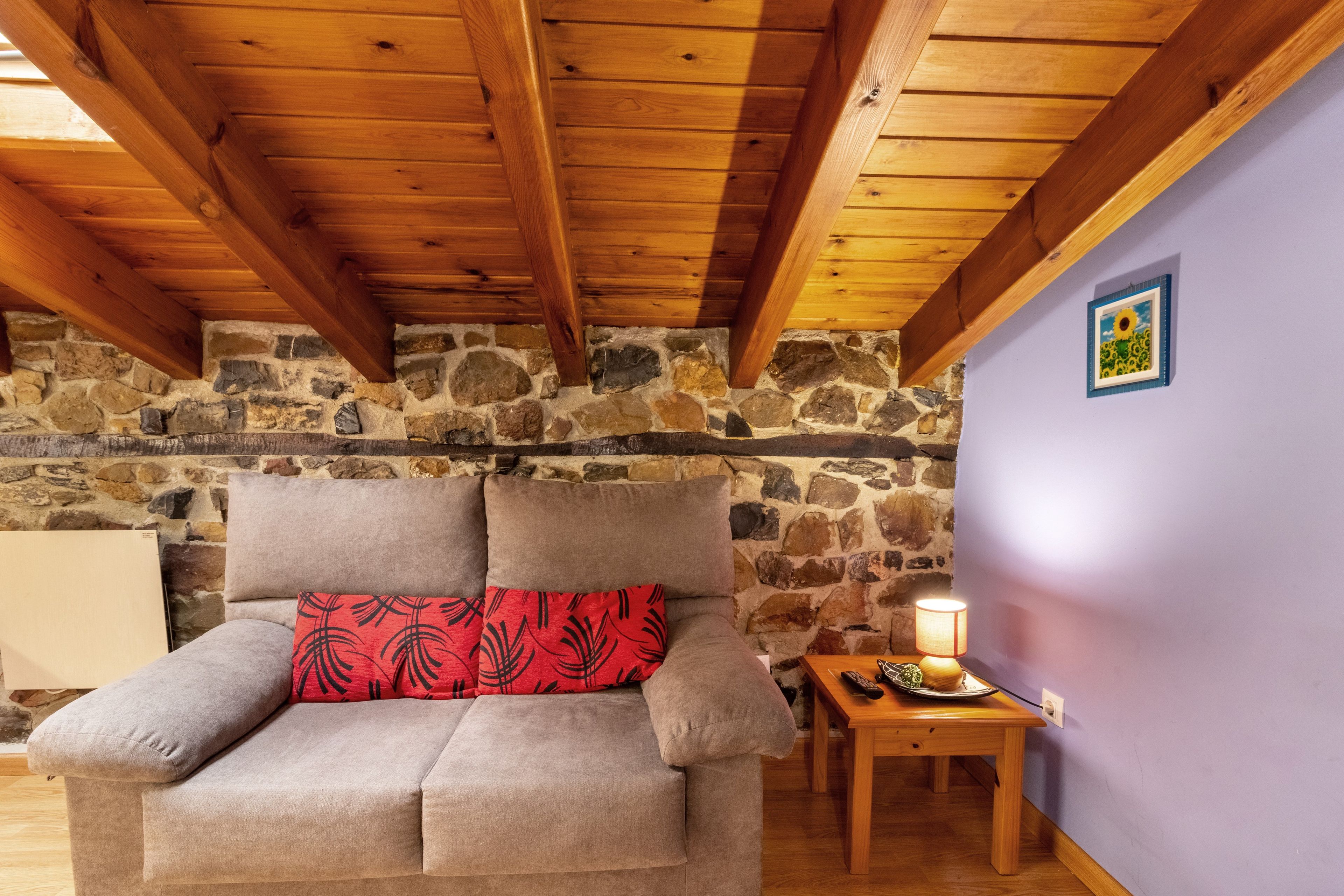 Una habitación acogedora con pared de piedra, techo de madera, un sillón con cojín rojo y una mesita con lámpara.