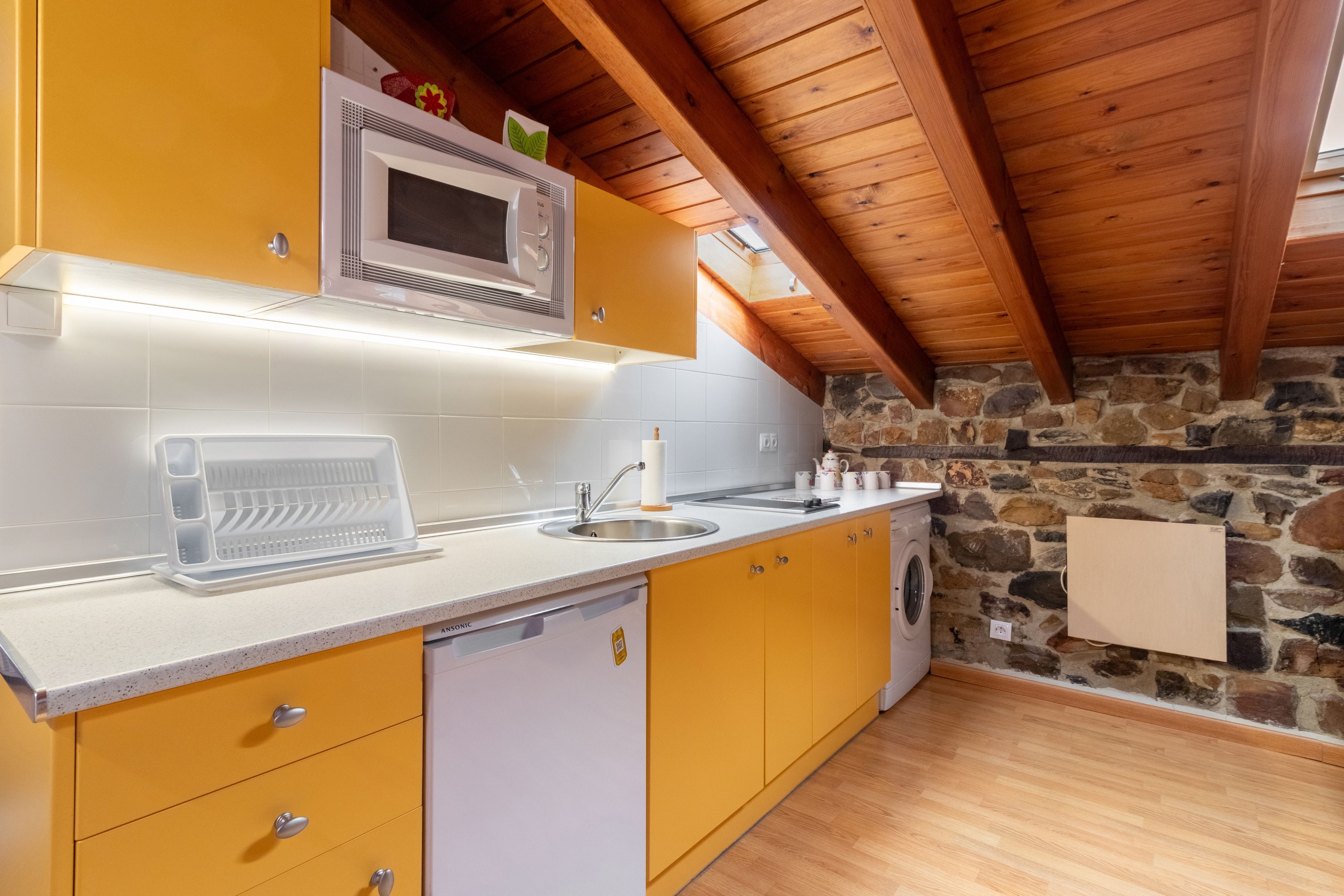 Una cocina con armarios amarillos, encimera blanca, microondas integrado, lavavajillas y pared de piedra.