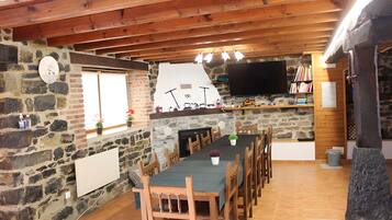 Un coin repas rustique avec un mur en pierre, des poutres en bois au plafond, un téléviseur à écran plat fixé au mur et une longue table entourée de chaises.