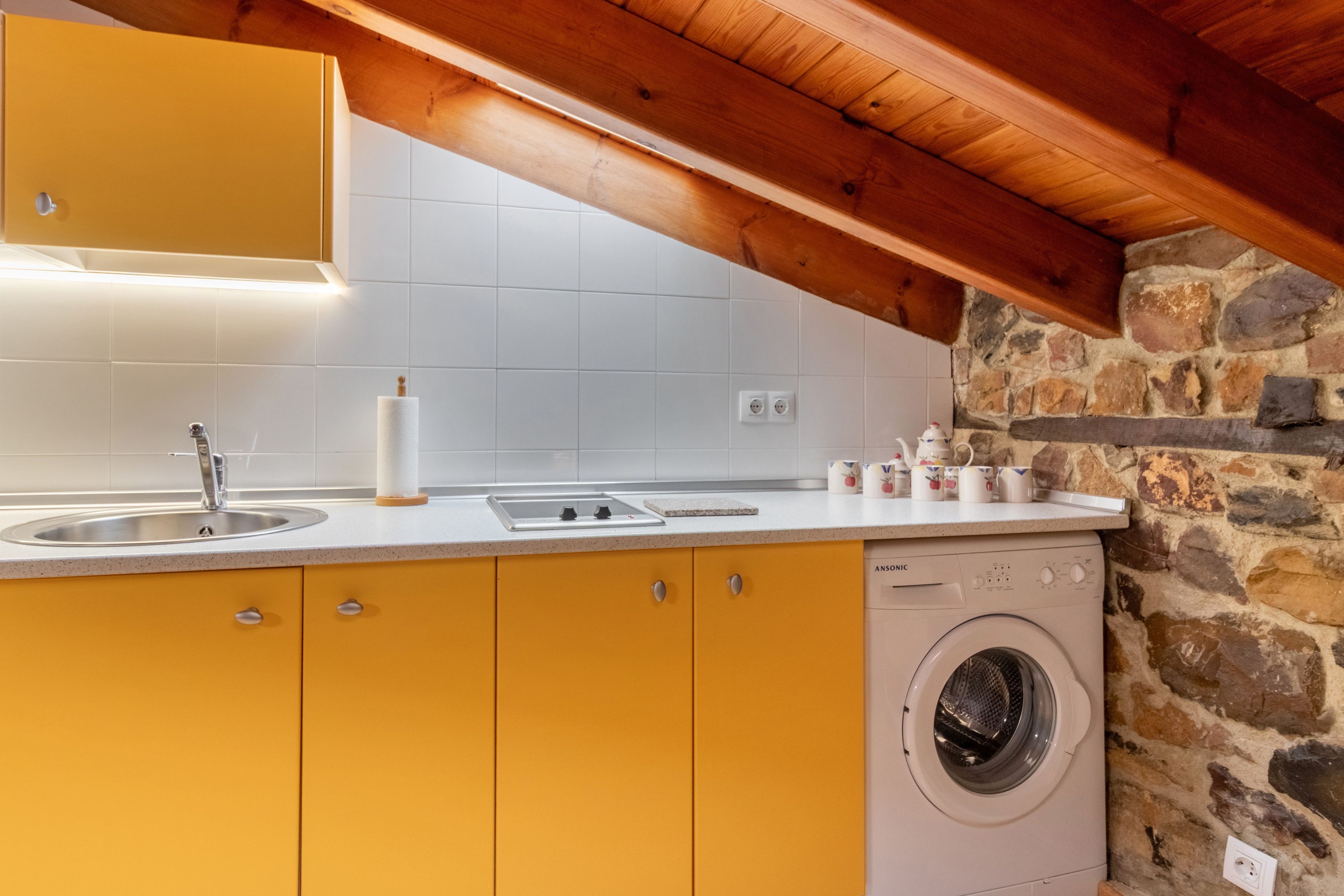 Une cuisine avec des meubles de couleur jaune, un lave-linge et un mur en pierre.