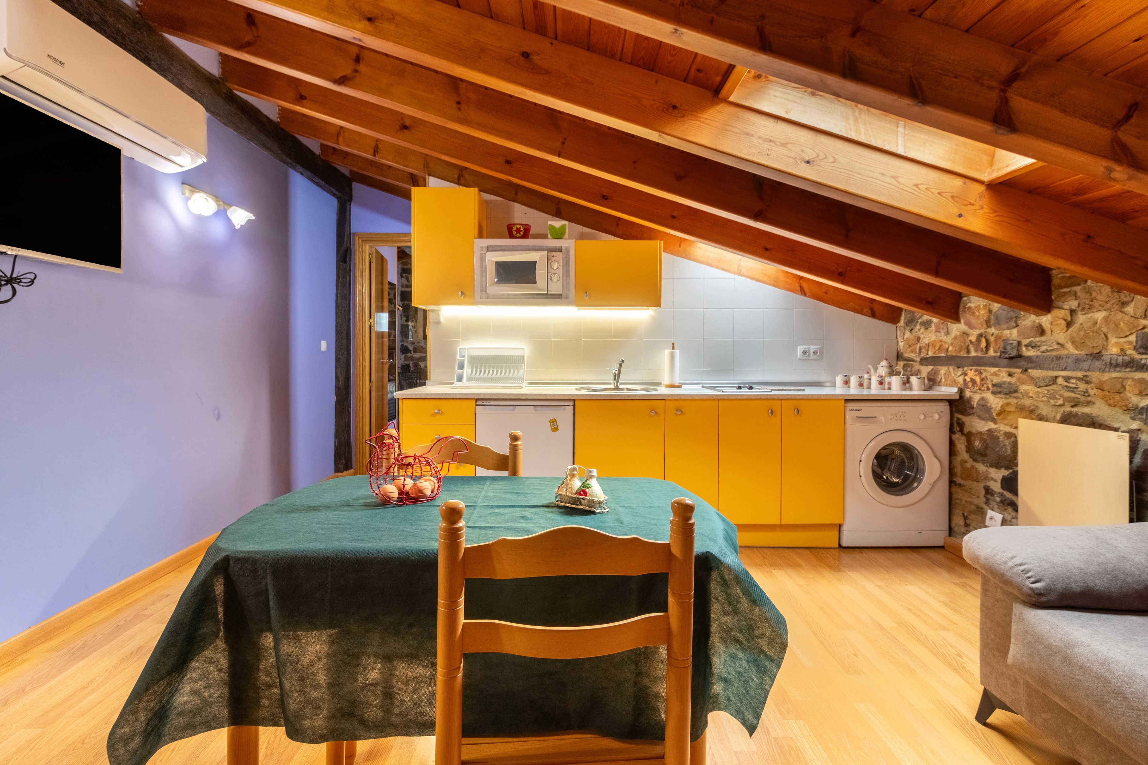 Une cuisine chaleureuse avec des poutres en bois, une table à manger recouverte d’une nappe verte et un lave-linge.
