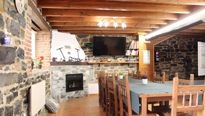 Un interior rústico con pared de piedra, techo de madera, chimenea, televisor y zona de comedor con sillas y mesa de madera.