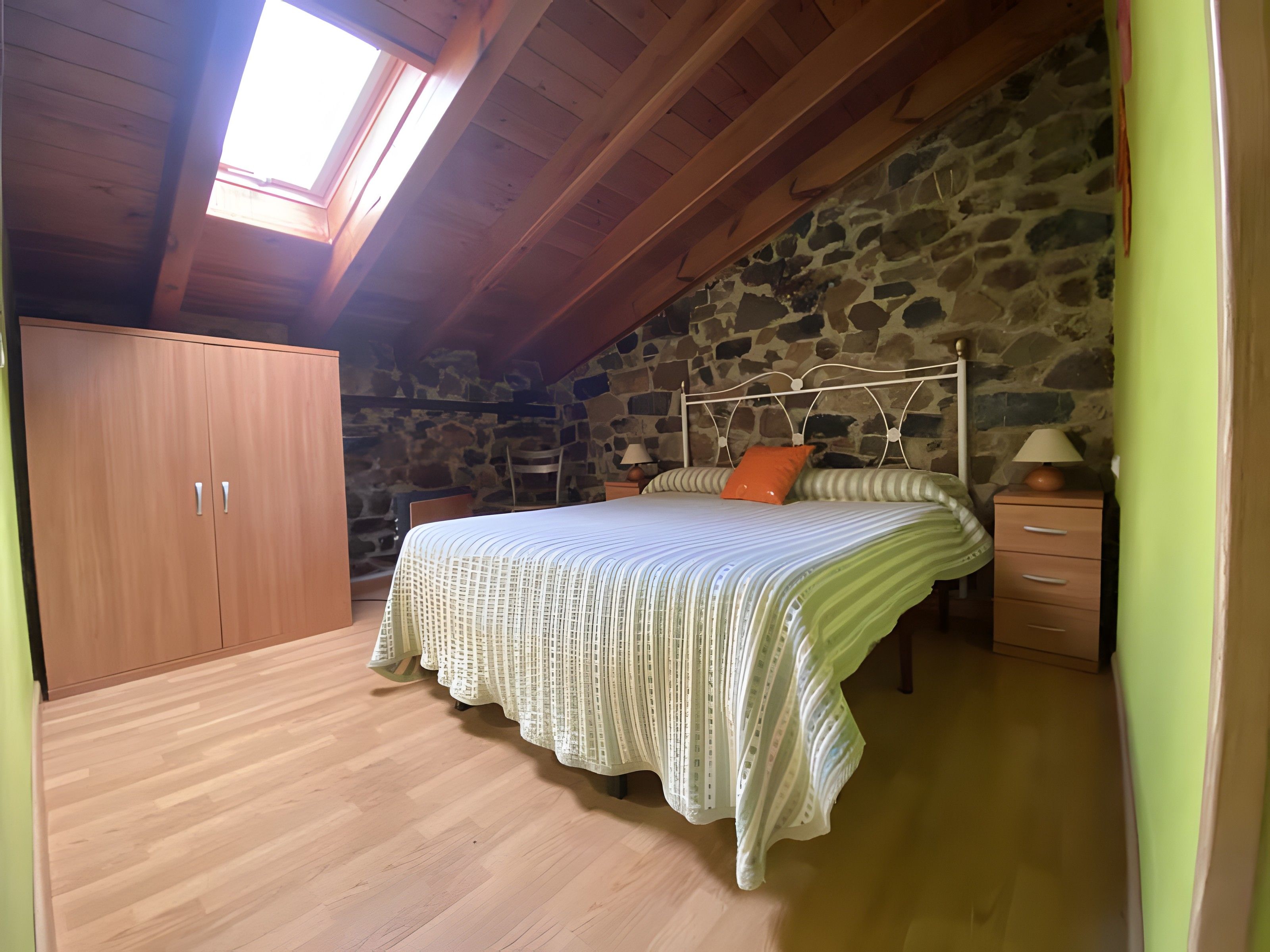 Een slaapkamer met een houten plafond, een stenen muur, een bed met gestreept beddengoed, een nachtkastje en een garderobe.