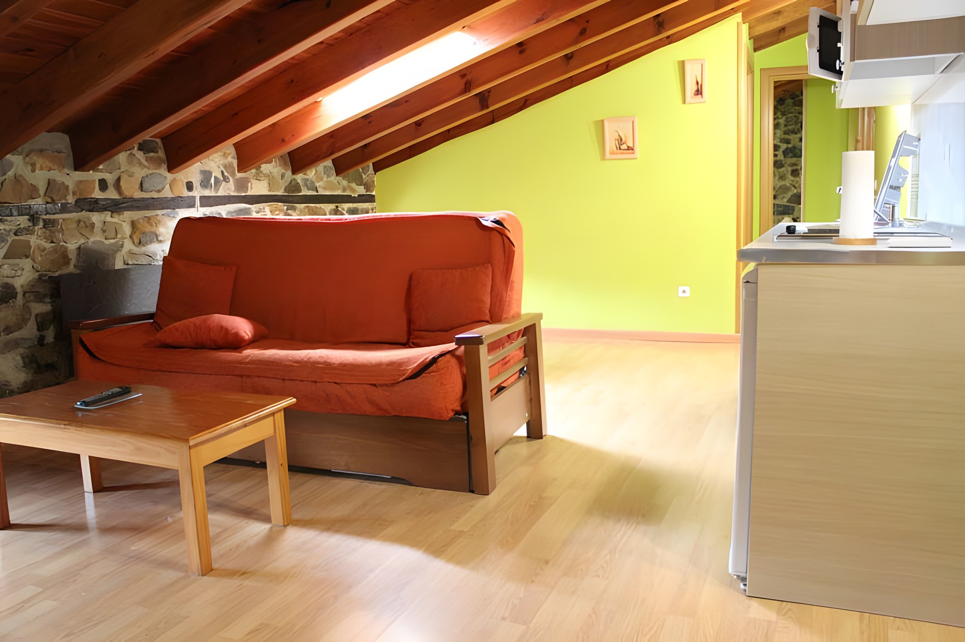 Una habitación acogedora con techo de madera, pared de piedra, un sofá, una mesita y una zona de cocina.