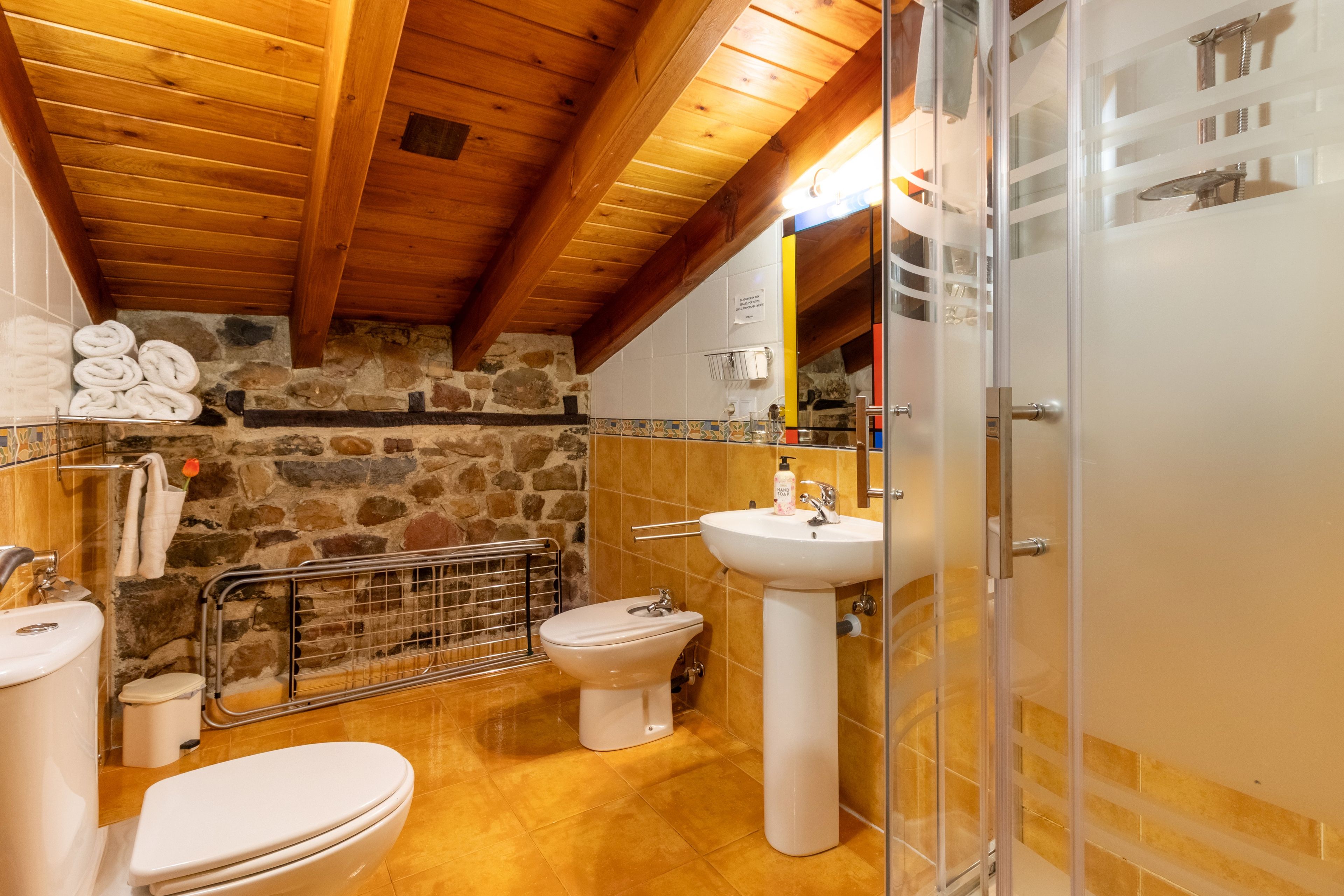 Un baño con pared de piedra, vigas de madera en el techo, un lavamanos blanco, inodoro y una cabina de ducha con paredes de vidrio.