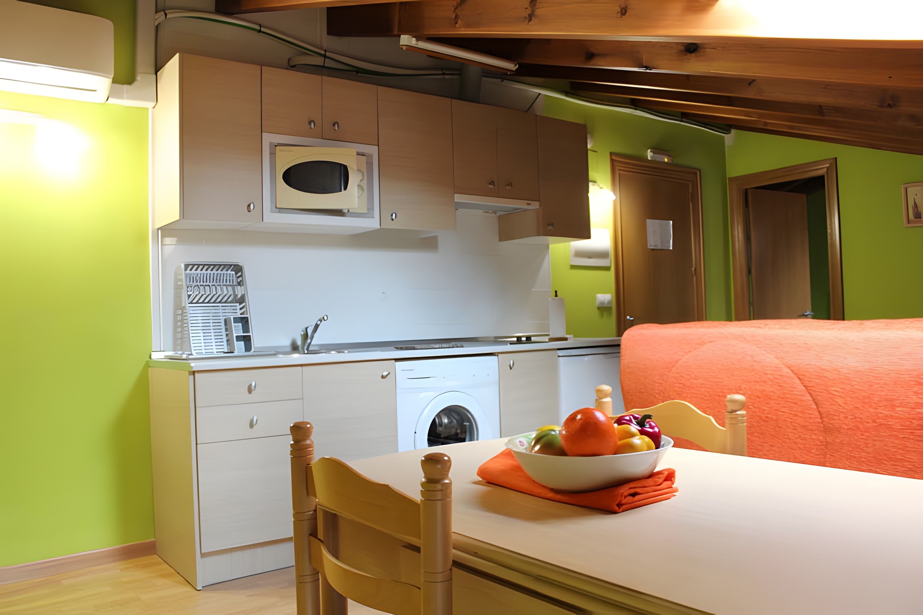 Cocina compacta con microondas, lavabo y lavadora. En primer plano, una mesa con un cuenco de fruta.