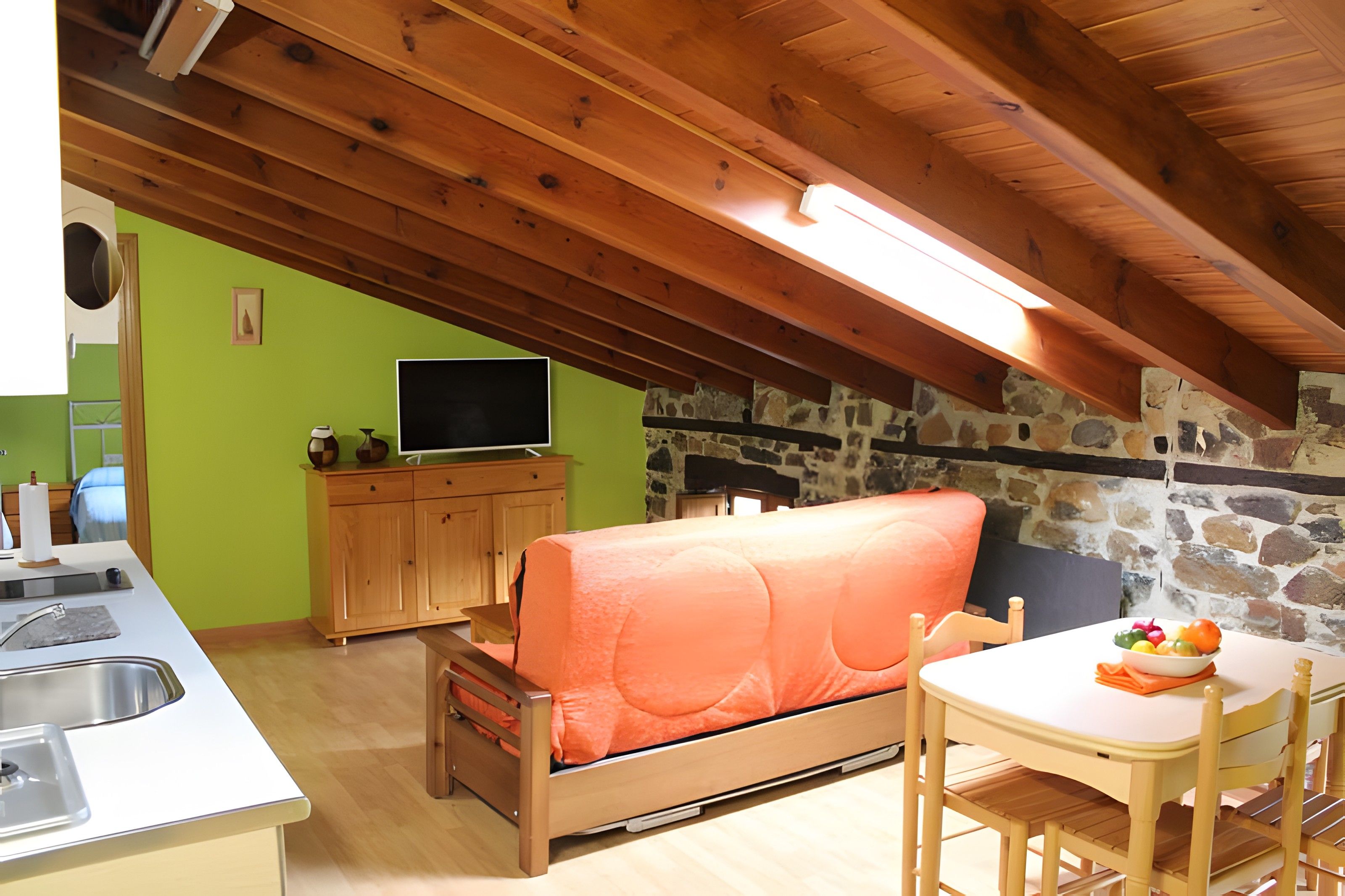 Een gezellig slaapkamer op zolder met een houten plafond, een stenen muur, een bed, een televisie en een eethoek met een tafel en stoelen.