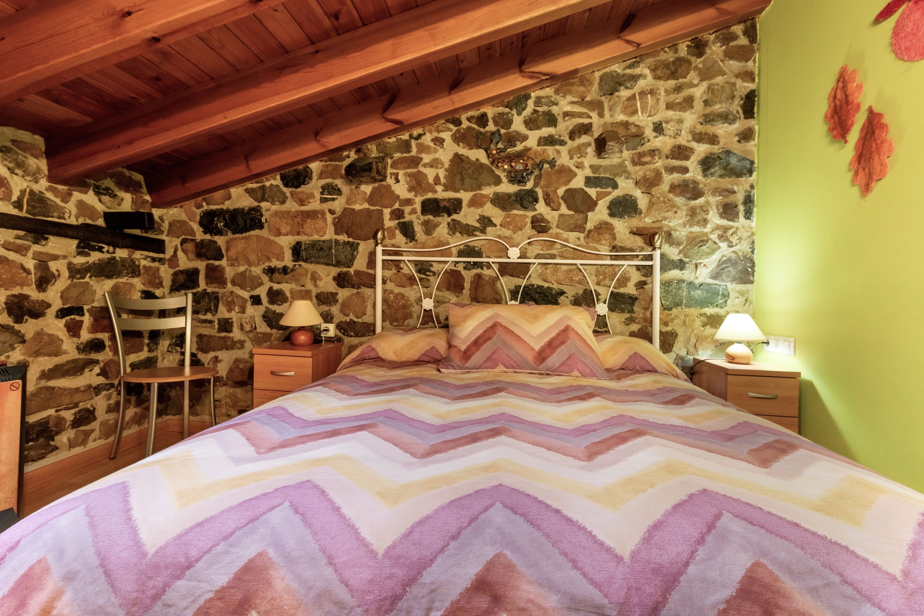 Un dormitorio con pared de piedra, techo de madera, cama con colcha estampada, mesita de noche con lámpara y una silla.